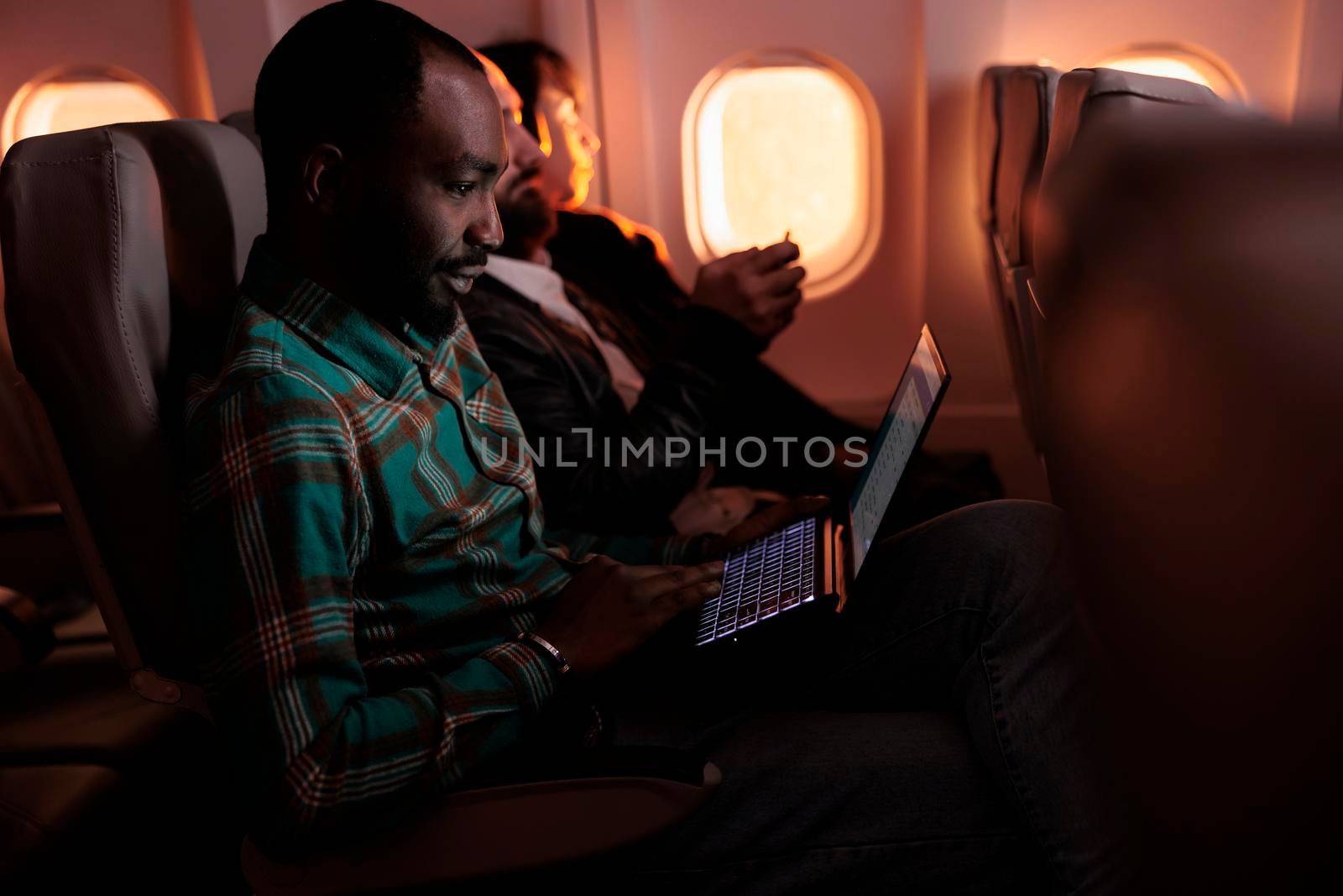Modern traveller browsing internet on laptop and flying abroad in airplane, using international airline to travel on business work trip. Young man working on computer and travelling during sunset.