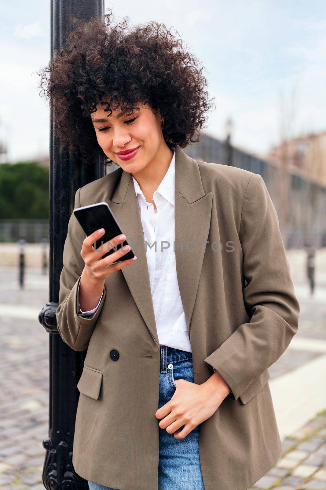 portrait of a smiling young latin woman checking her phone leaning against a city lamppost, technology and communication concept