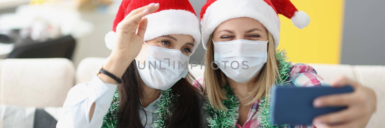 Portrait of cute best friends taking fun selfie in face masks on smartphone and laugh. Home party before christmas. New year, pandemic, celebration concept