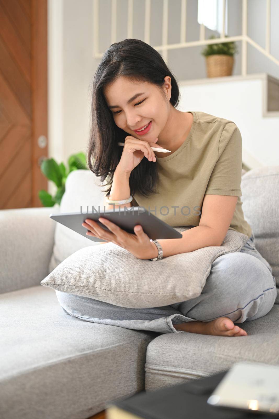 Smiling young woman reading ebook on digital tablet while relaxing in comfortable couch at home.