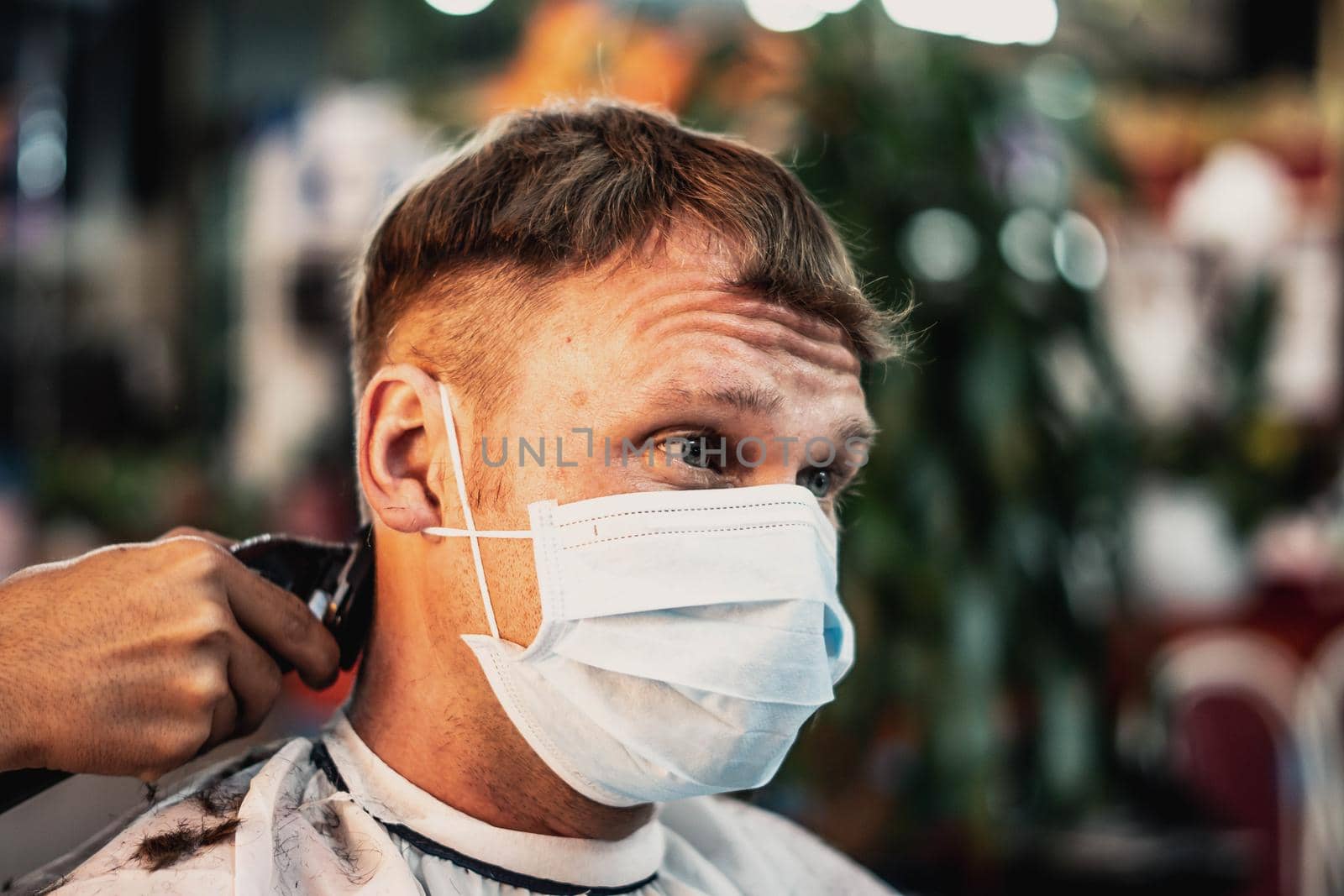 Lifestyle photo barbershop. Young man customer wears face mask, surprised dissatisfied look, wrinkled forehead. Hairdresser male hand work comb tool electric clipper. Short hair locks cut.