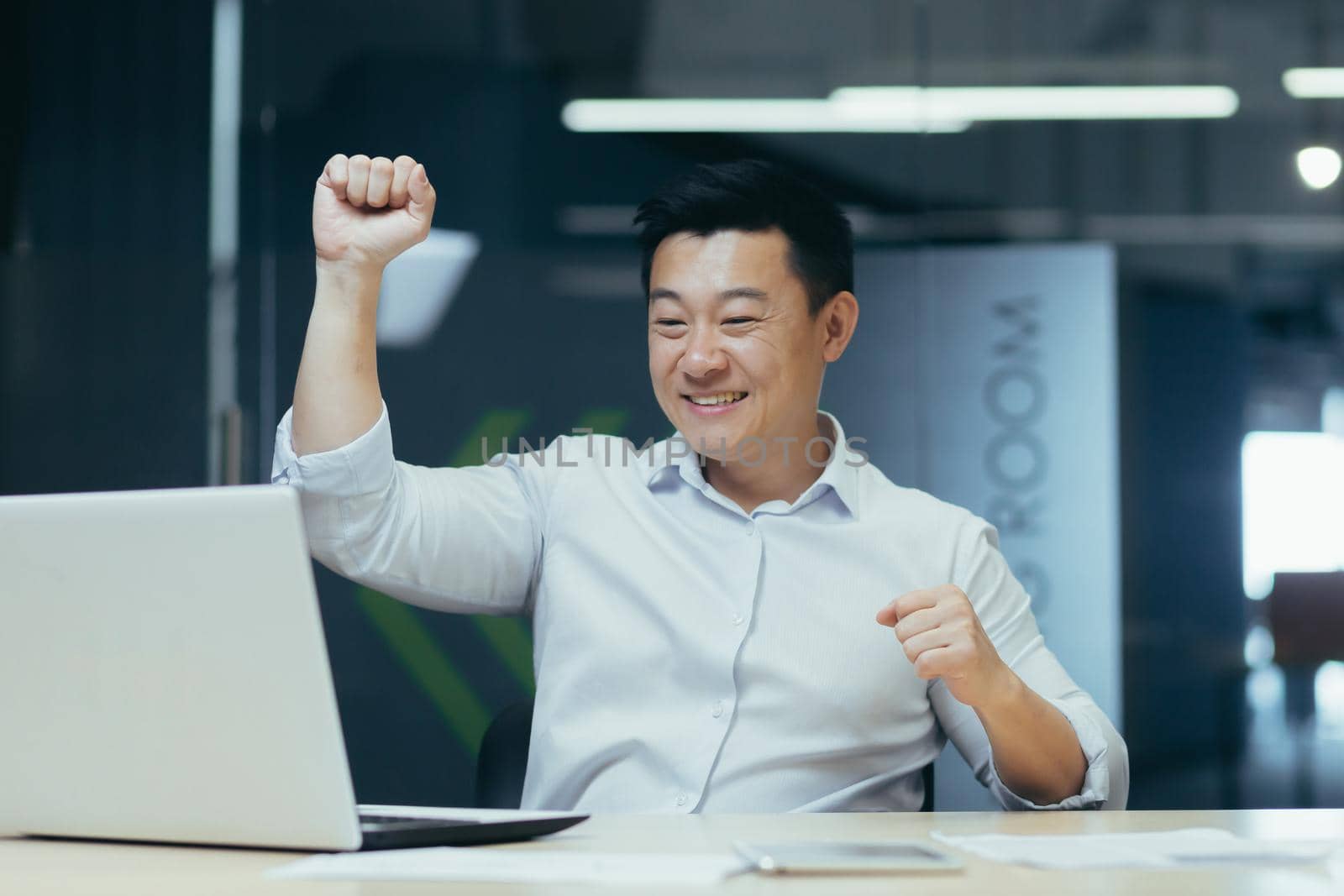 Cheerful and successful asian businessman dancing in office celebrating triumph and victory sitting at desk working with laptop