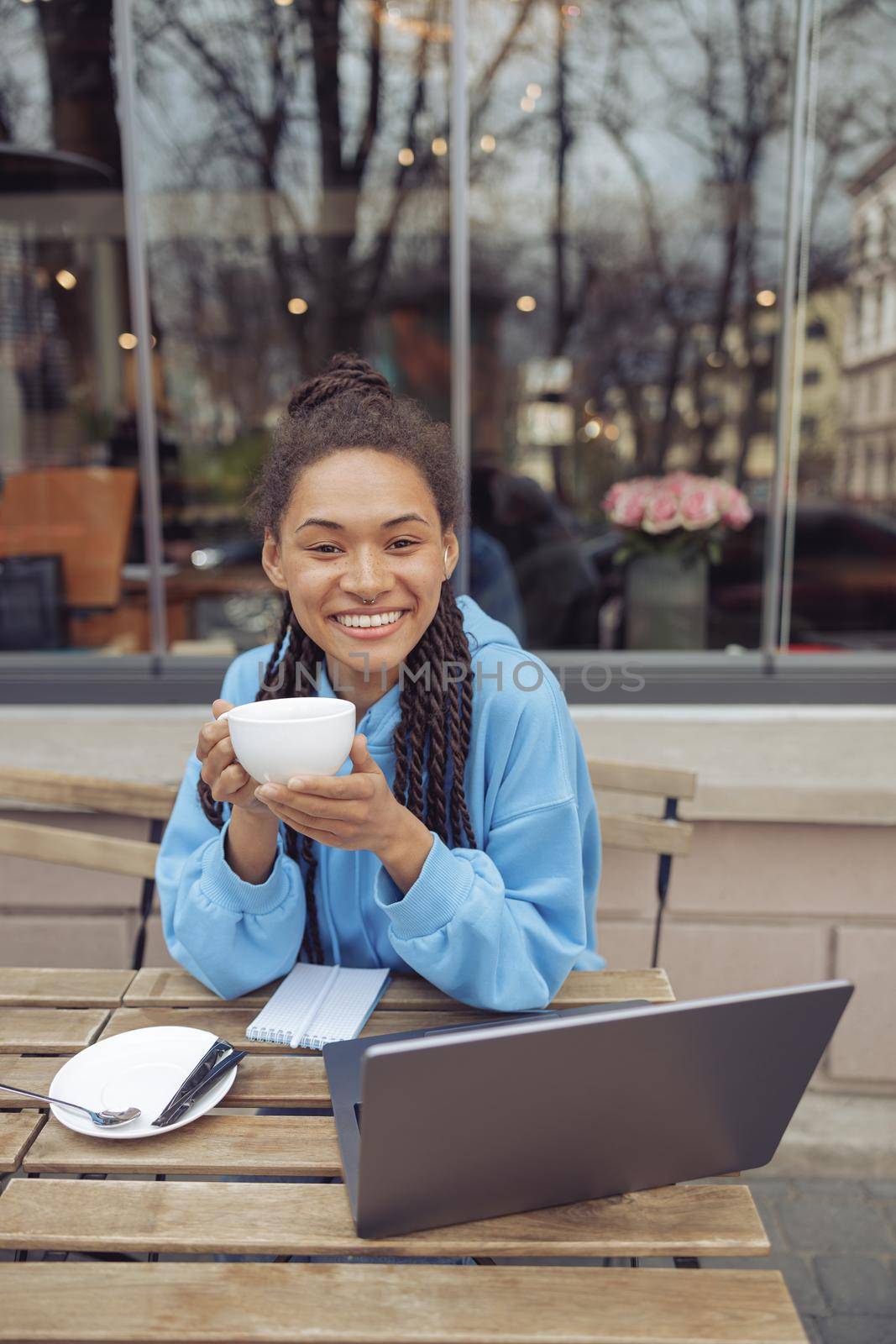 Portrait of happy young pretty woman smiling while sitting with coffee at laptop outside. Vertical.