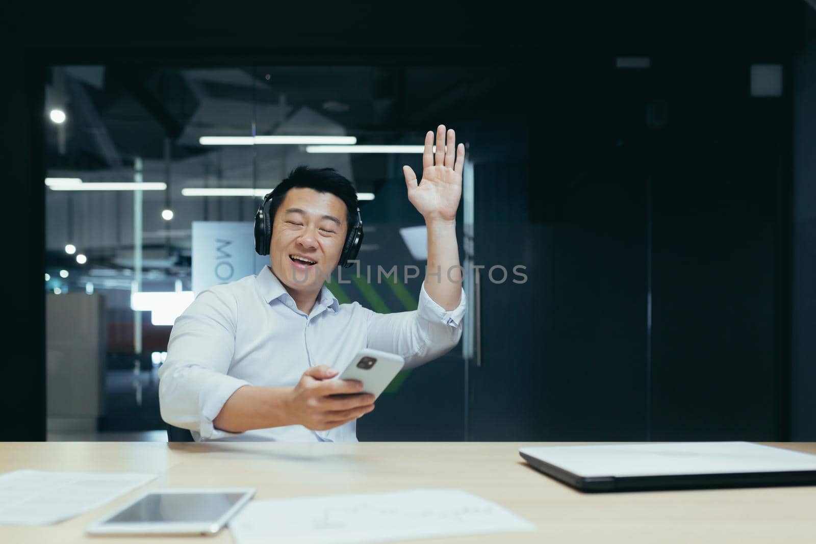 Businessman in office resting during work break, listening to music in big black headphones, Asian man using music app on phone