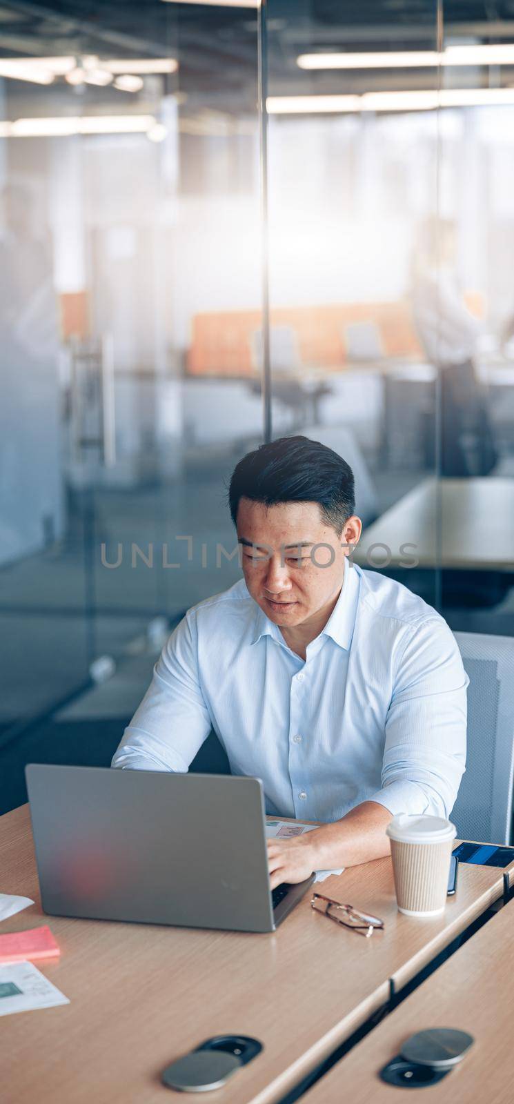 Focused asian businessman working on laptop at her workplace with colleagues in the background.