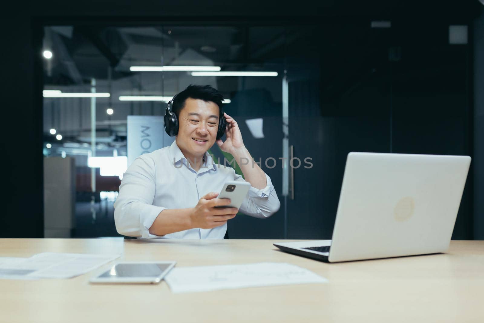 Businessman in office resting during work break, listening to music in big black headphones, Asian man using music app on phone