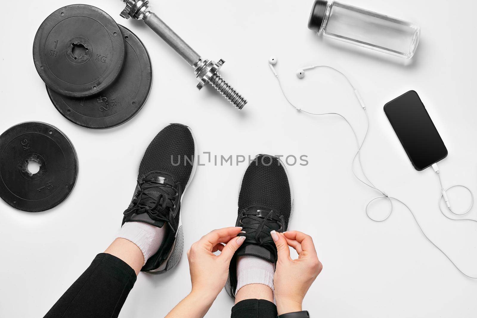 Young woman in sporting leggings laces sneakers, preparing for training. Accessories for sports, bottle of water, smartphone, headphones, fitness bracelet, dumbbells on white background flat lay top view. Fitness concept