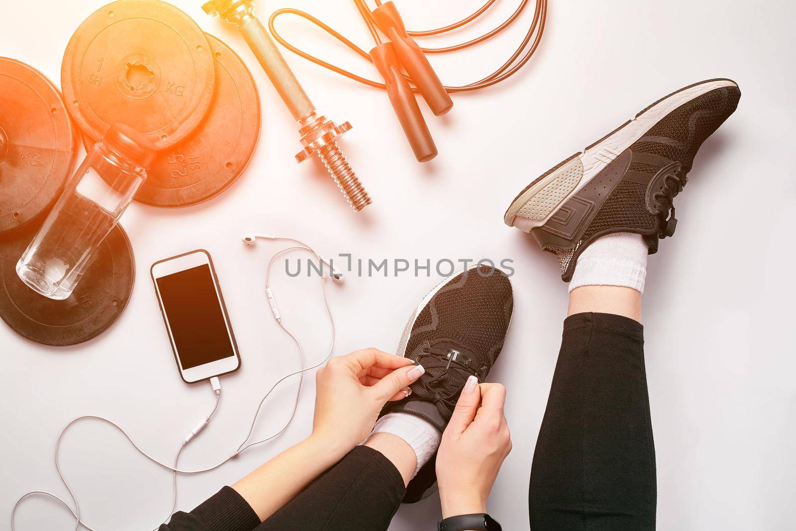 Young woman in sporting leggings laces sneakers, preparing for training. Accessories for sports, bottle of water, smartphone, headphones, fitness bracelet, dumbbells on white background flat lay top view. Fitness concept. Sun flare