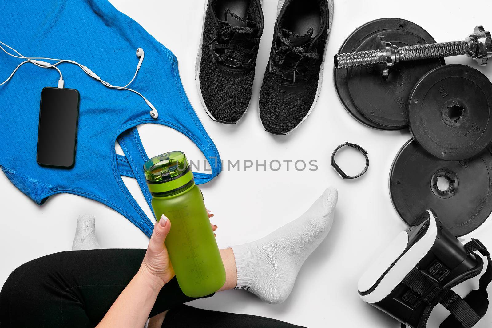 Flat lay gym equipment such as dumbbells, bottle of waters, smartphone with headphone, fitness bracelet and human legs in socks on white background. Fitness concept
