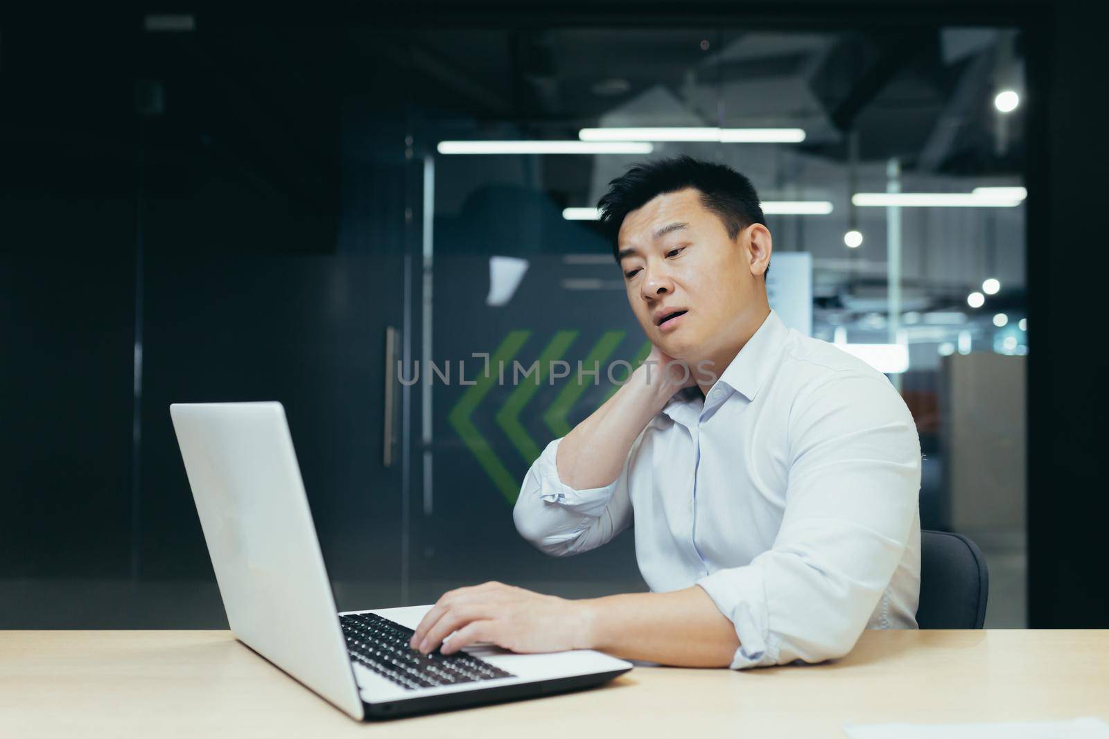 Overtired asian worker in office, having severe neck pain, massaging with hand, businessman working in office with laptop