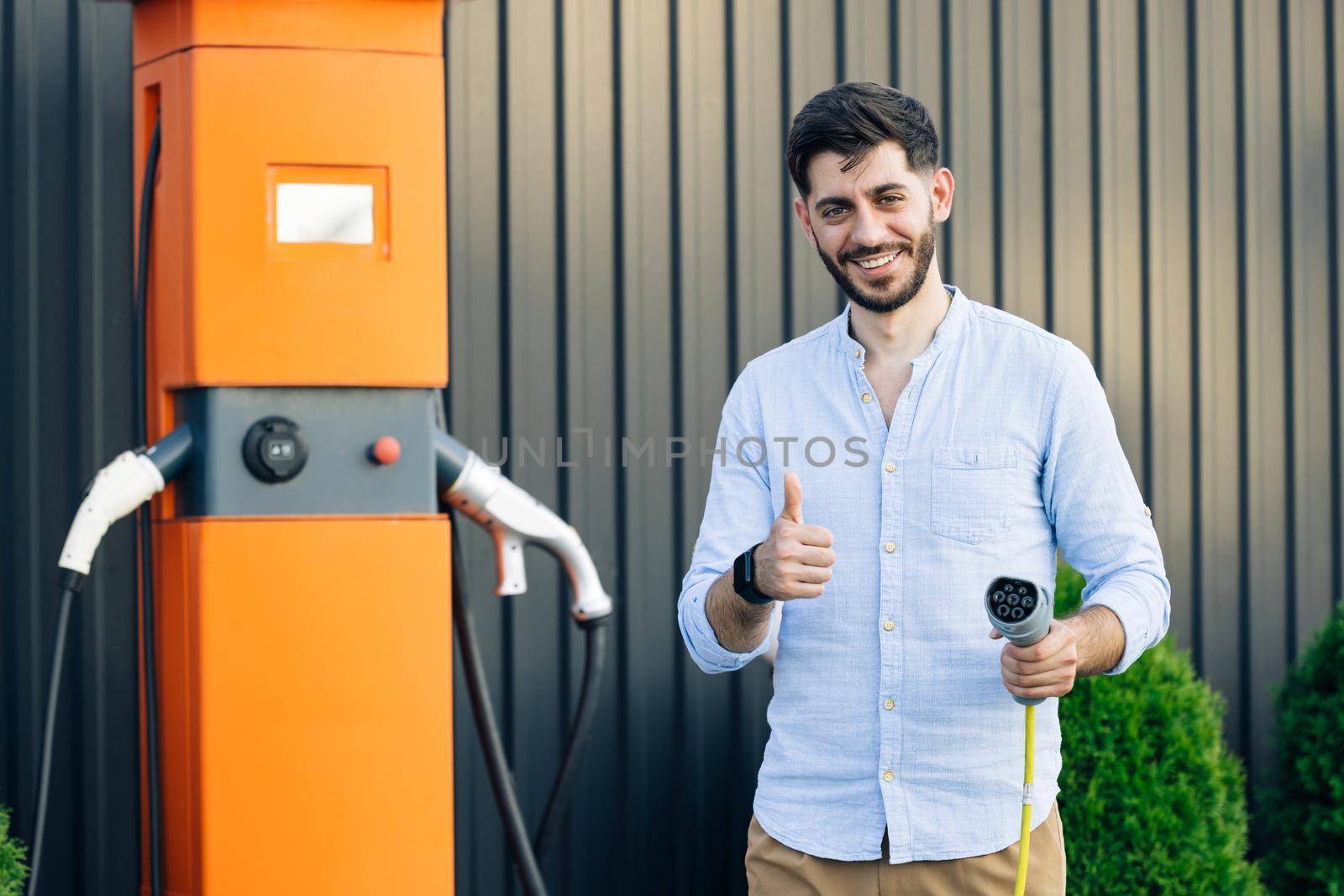 Caucasian bearded hipster man standing near electric charging station looking at camera and showing thumb up. Male holds a charging cable in his hands. Eco-friendly modern green transport.