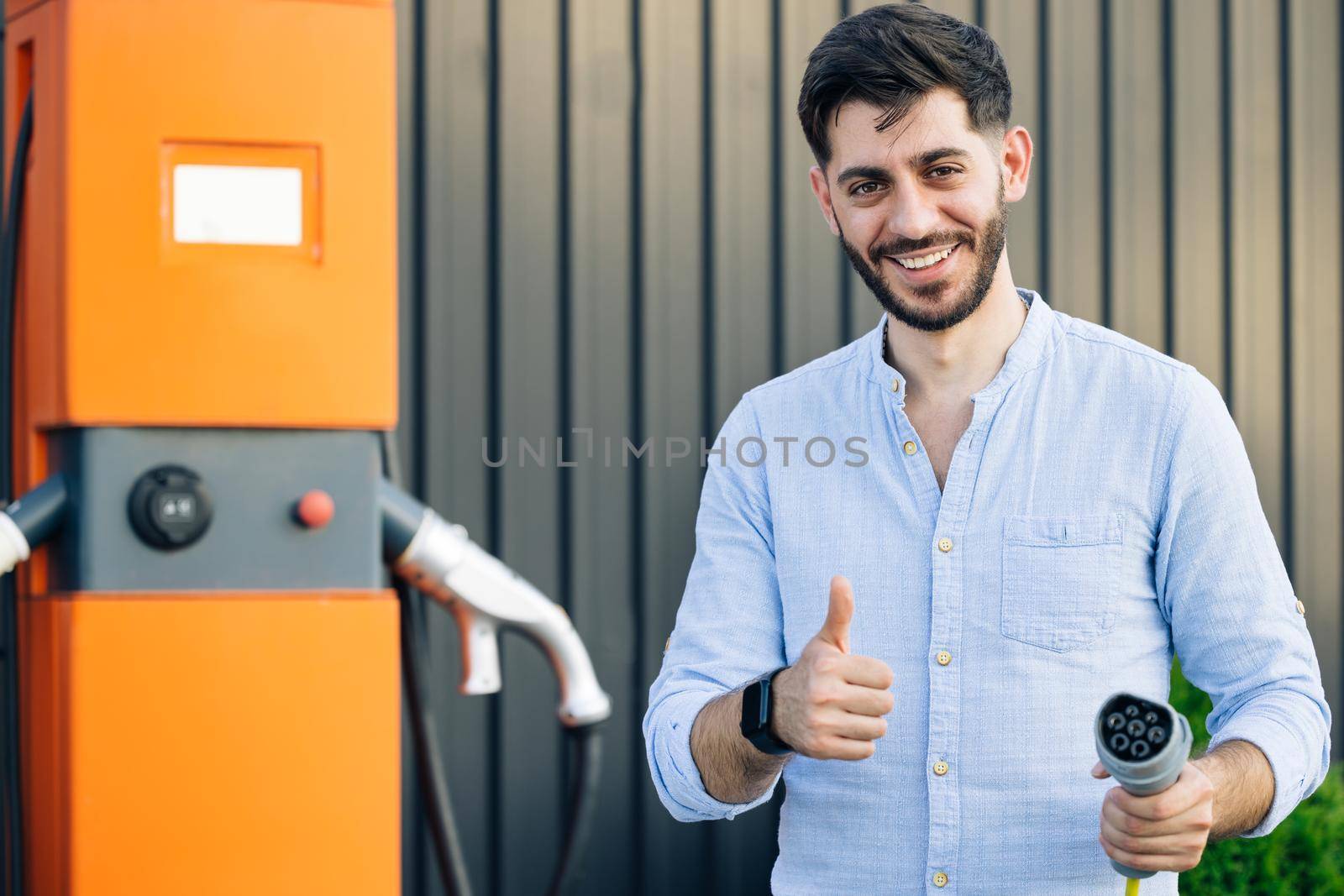 Male holds a charging cable in his hands. Caucasian bearded hipster man standing near electric charging station looking at camera and showing thumb up. Eco-friendly modern green transport by uflypro