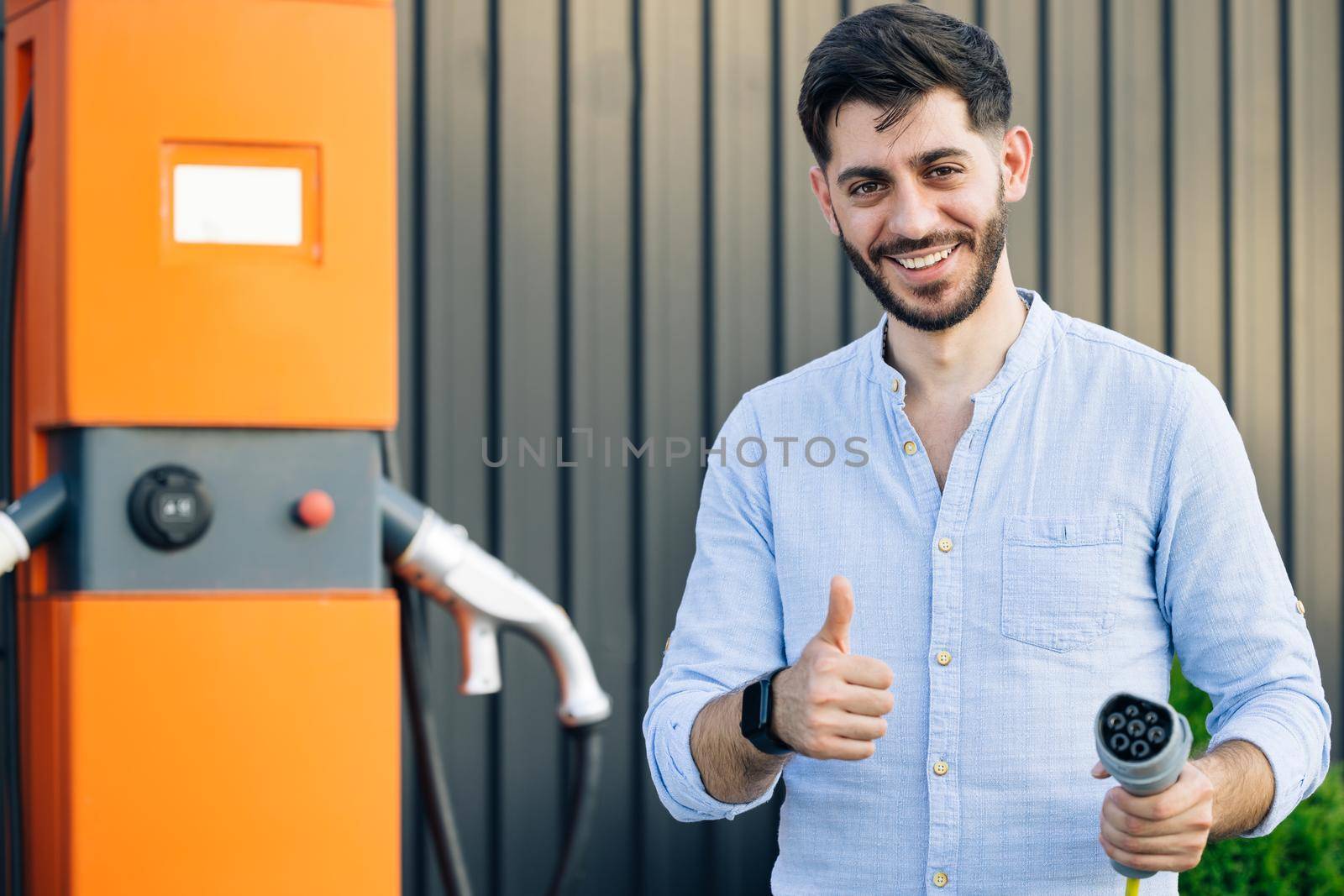 Male holds a charging cable in his hands. Caucasian bearded hipster man standing near electric charging station looking at camera and showing thumb up. Eco-friendly modern green transport.