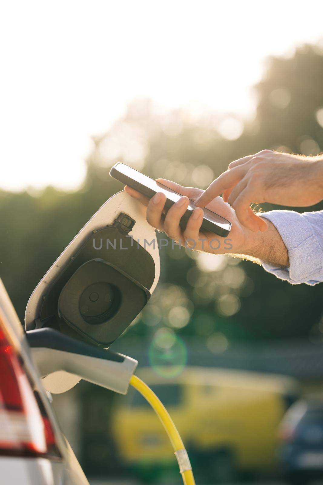 Man charging electric car at charging station using smart phone app. Unrecognizable male unplugging electric car from charging station. Man is unplugging in power cord to an electric car at sunset by uflypro