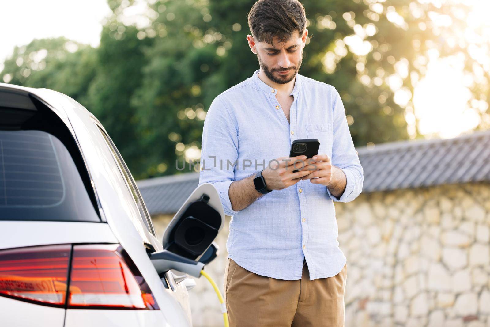 Caucasian bearded man using smart phone and waiting power supply connect to electric vehicles for charging the battery in car. Plug charging an Electric car from charging station by uflypro