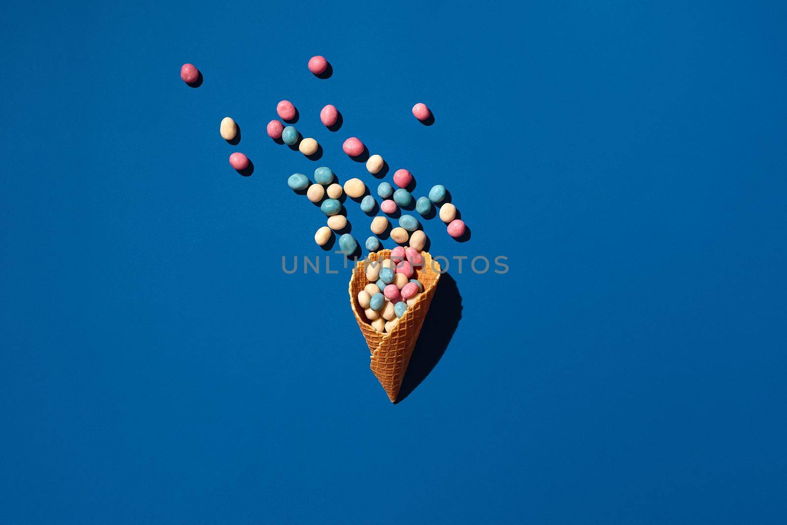 Flat lay photo of a heap of candy dragee that is sprinkled from a waffle cone on blue background from above