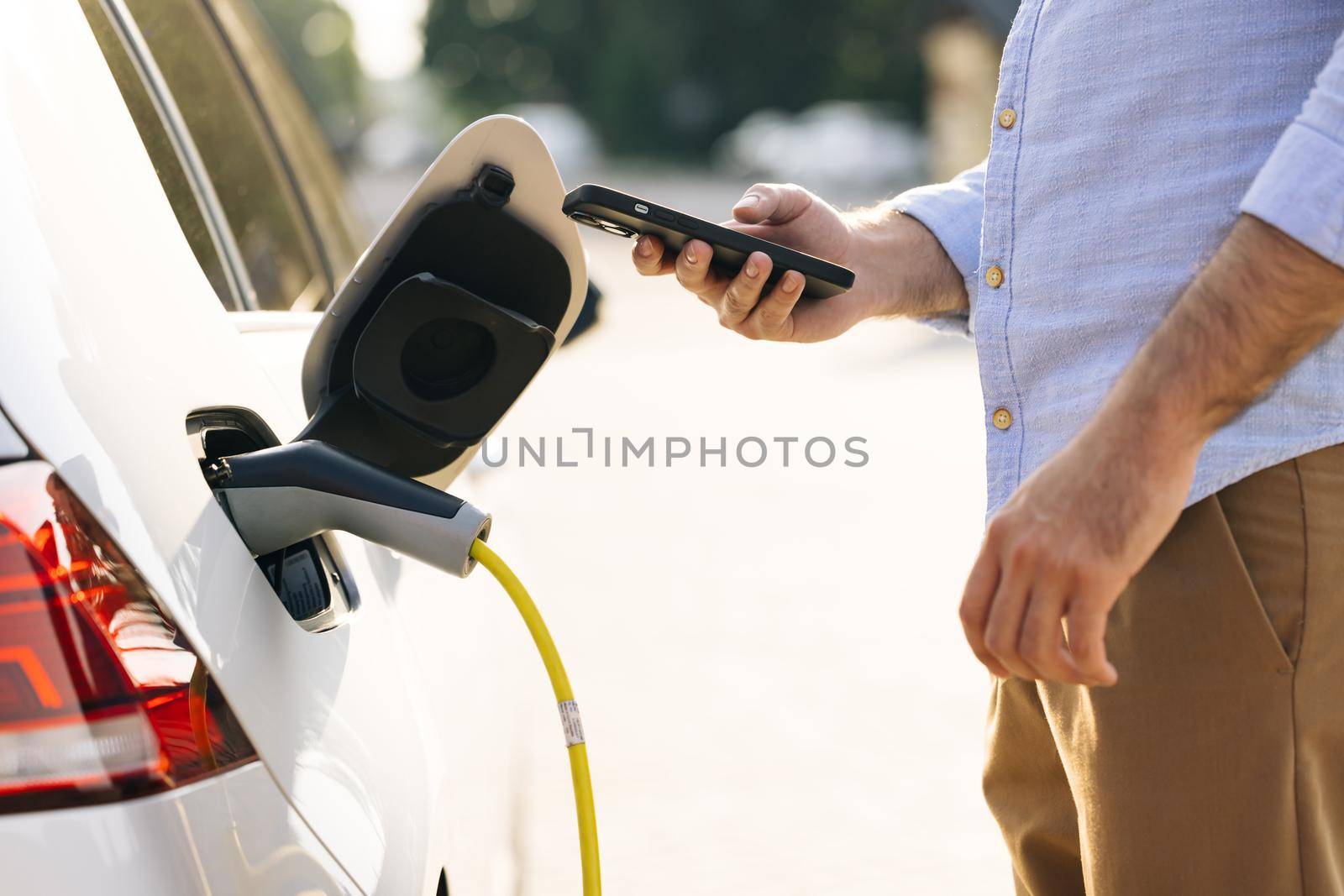Unrecognizable man plugging electric car from charging station. Male plugging in power cord to electric car using app on smartphone. Businessman charging electric car at outdoor charging station