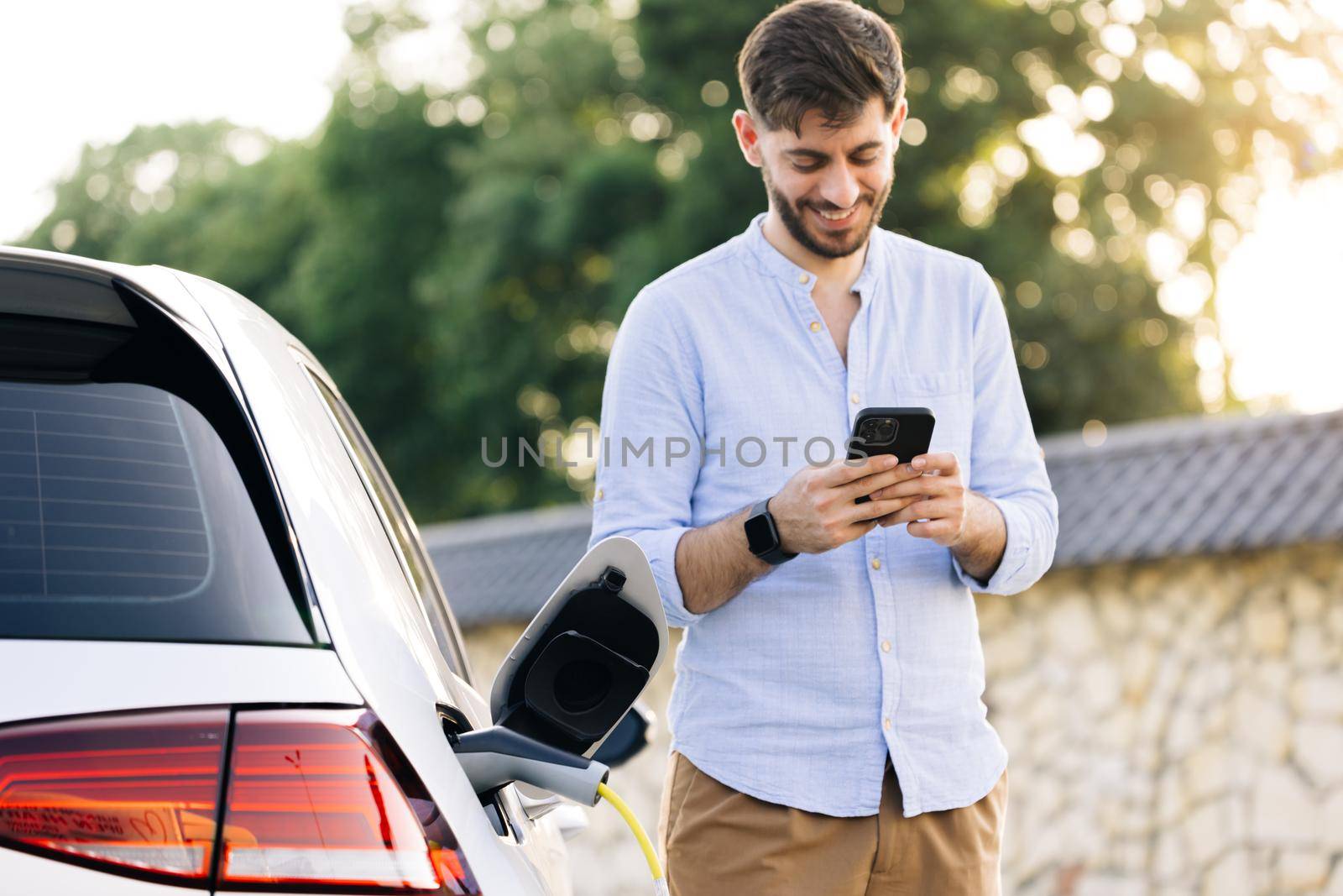 Bearded caucasian man in a blue shirt standing near an electric car that is charging and making time adjustments on a smartphone. Plug charging an Electric car from charging station.
