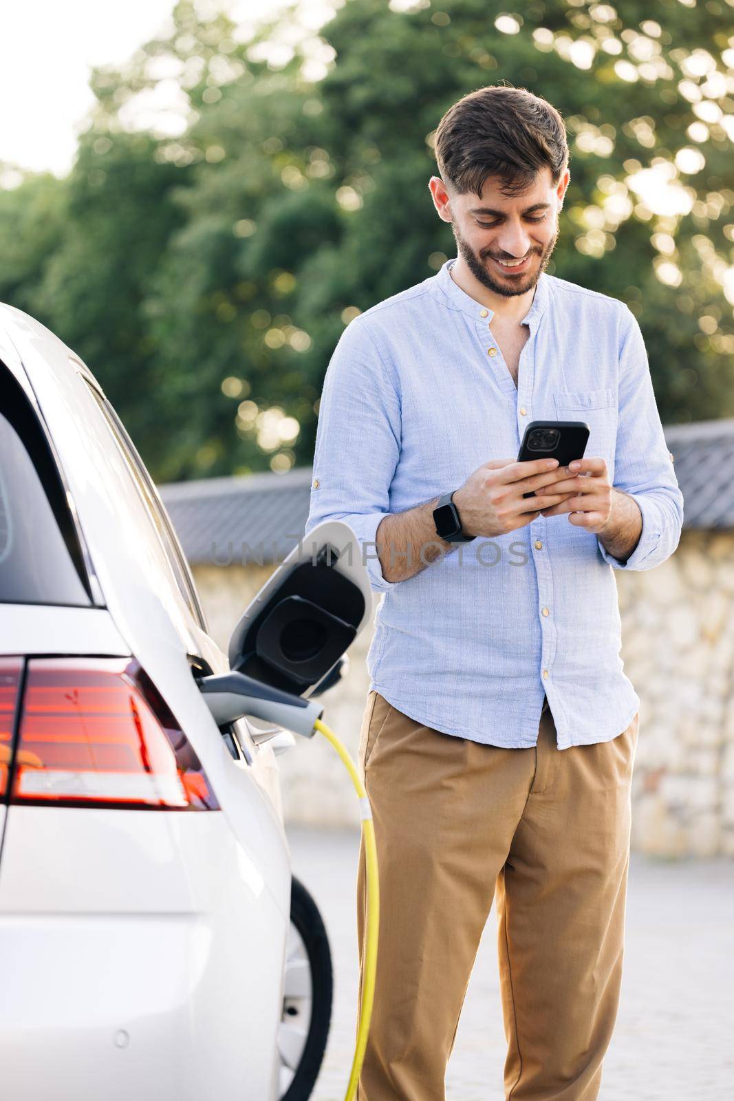 Bearded man using smart phone and waiting power supply connect to electric vehicles for charging the battery in car. Plug charging an Electric car from charging station.