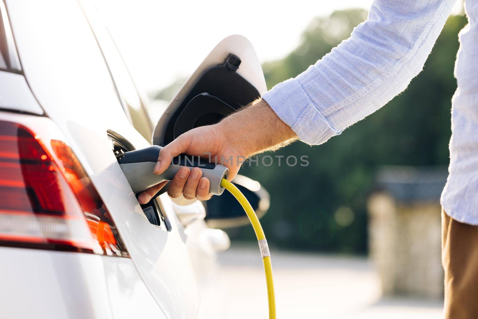 The driver of the electric car inserts the electrical connector to charge the batteries. Unrecognizable man attaching power cable to electric car. Electric vehicle Recharging battery charging port by uflypro