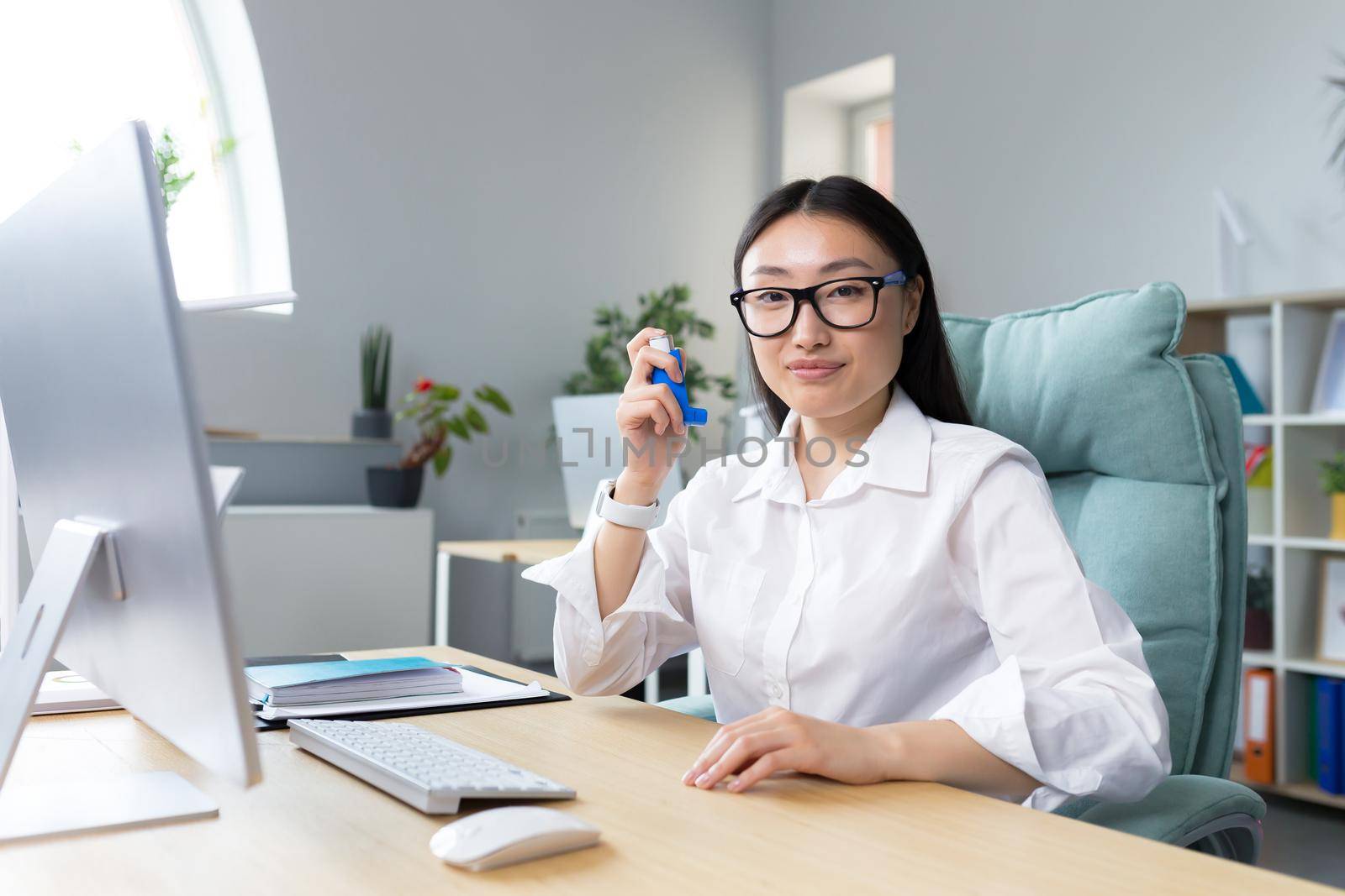 Allergy, feeling unwell at the workplace. A young Asian woman in glasses holds an inhaler in her hand, has allergies, asthma. Sitting in the office at the desk, looking at the camera.
