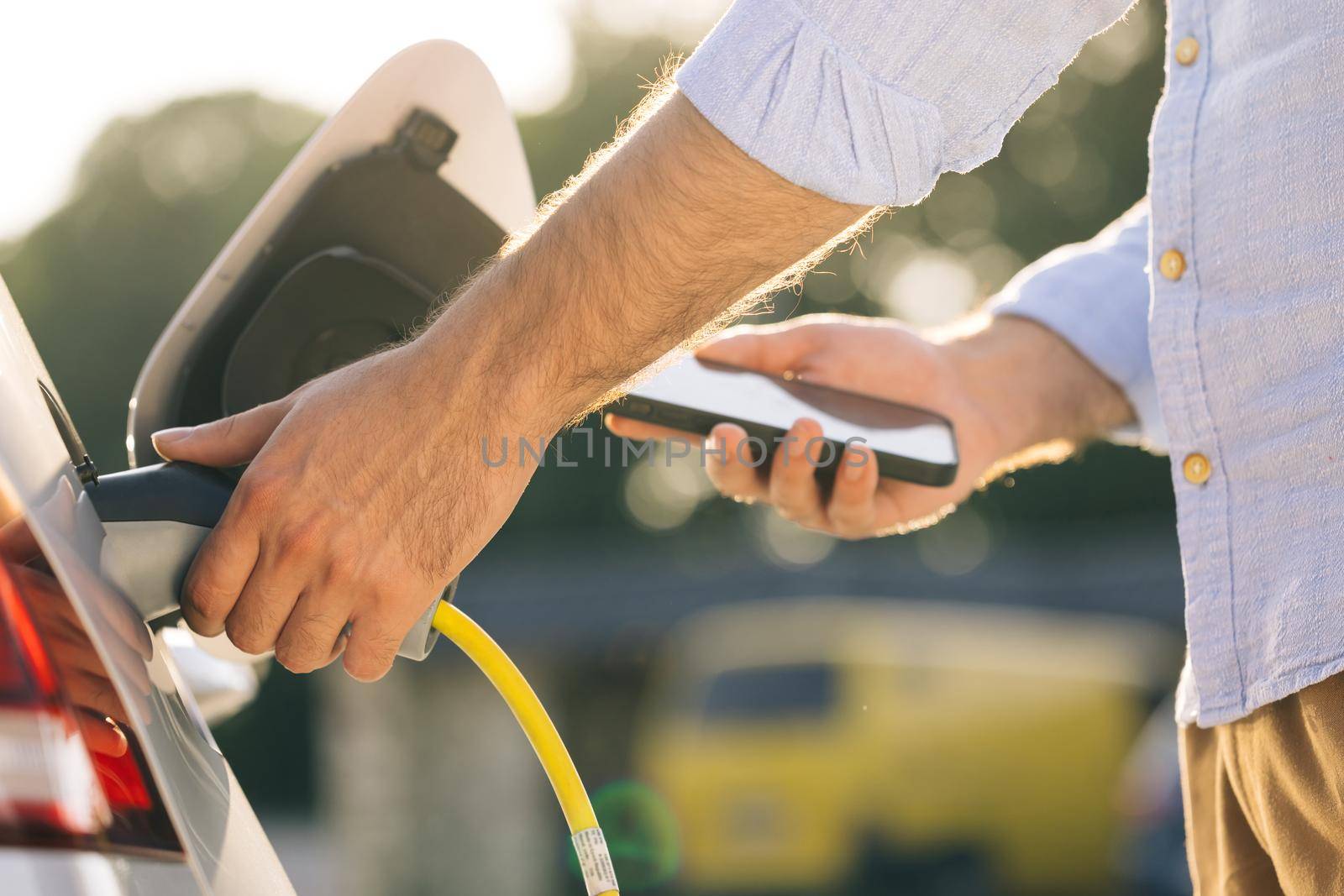 Man plugging electric car from charging station. Male attaching in power cord to electric car using app on smartphone. Businessman charging electric car on charging station at sunset