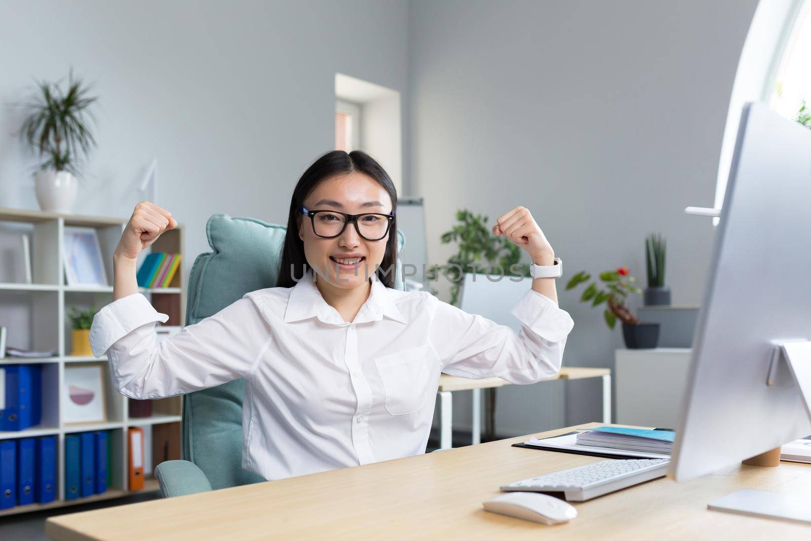 Portrait of a young beautiful Asian business woman who shows with her own hands that she is strong and successful. Sitting at the desk in the office wearing a white shirt and glasses.