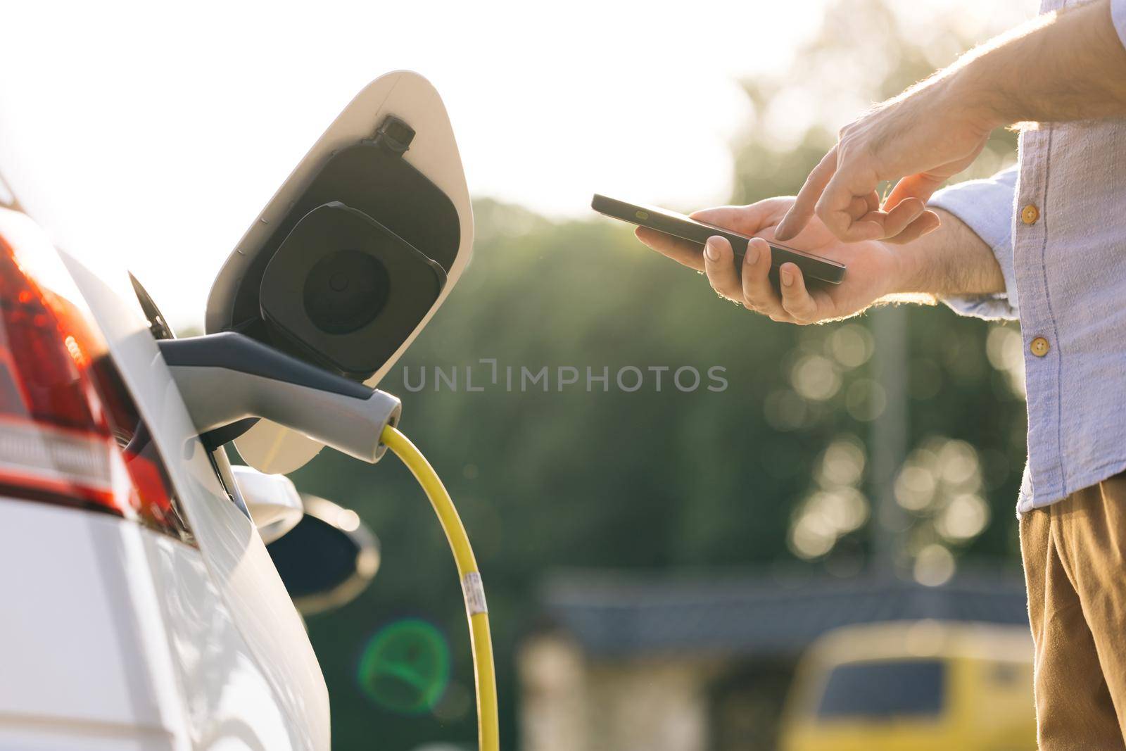 Businessman charging electric car at outdoor charging station. Unrecognizable man plugging electric car from charging station. Male plugging in power cord to electric car using app on smartphone.