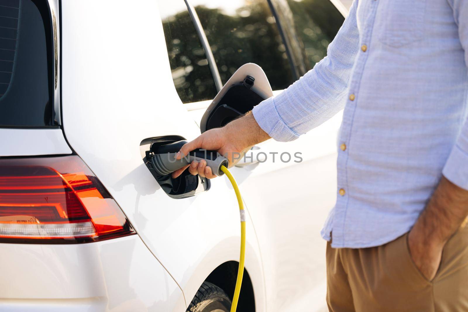 Man holding power supply cable at electric vehicle charging station. Hansome guy holding plug of the charger, while car is charging at the charging station by uflypro