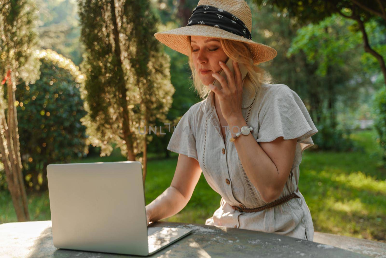 a beautiful mature woman in hat works on a computer at a white table in nature and spends her day productively.