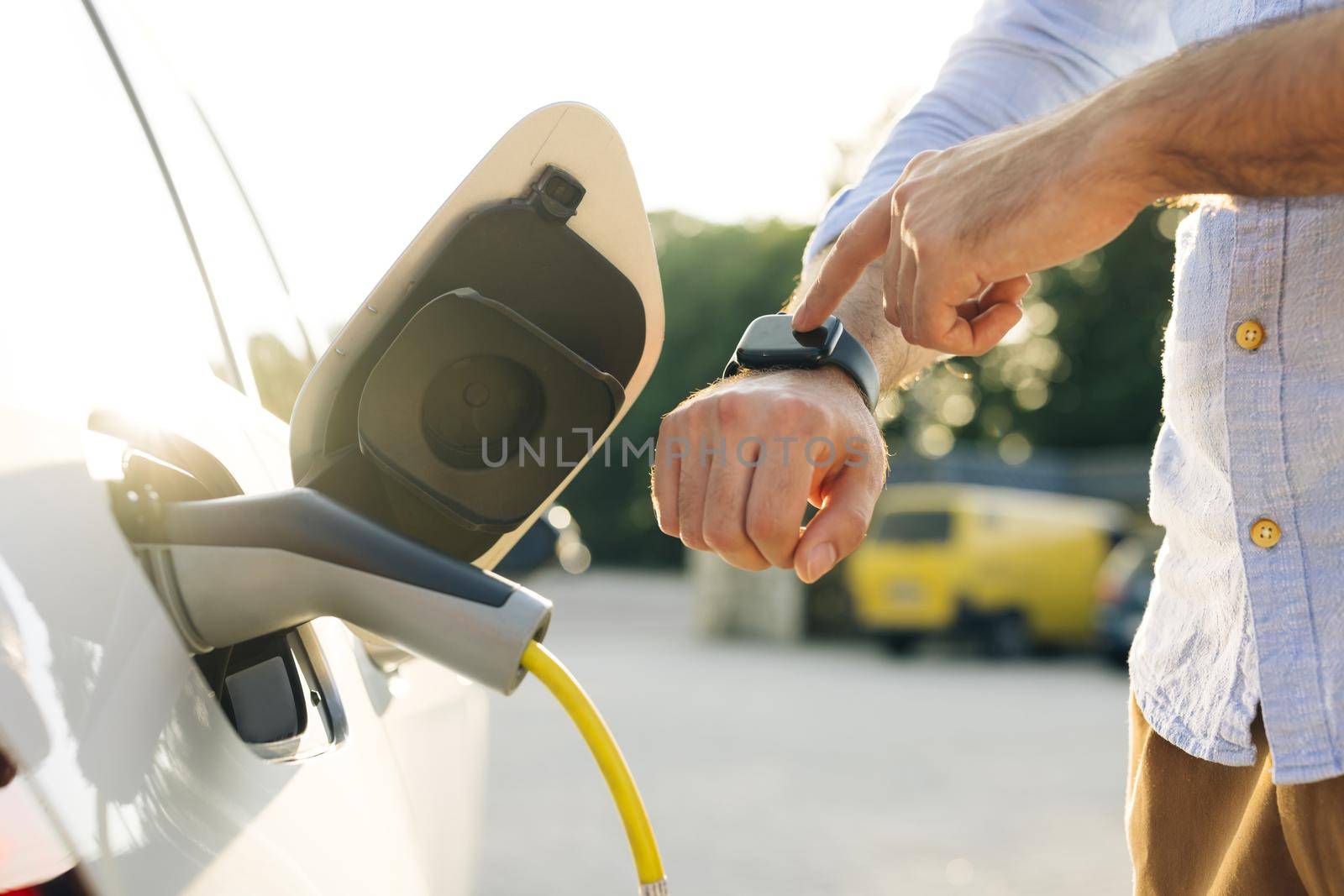 Businessman plugging in charging cable to to electric vehicle. Male hand inserts power connector into EV car and charges batteries, uses smartwatch for activates start charging