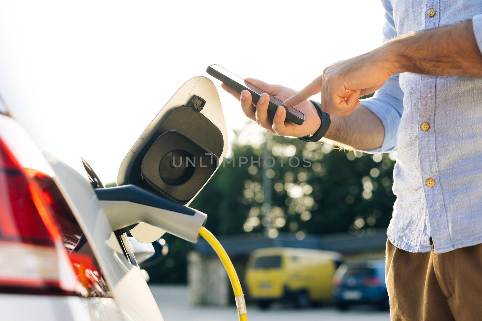 Male attaching in power cord to electric car using app on smartphone. Man plugging electric car from charging station. Businessman charging electric car on charging station at sunset