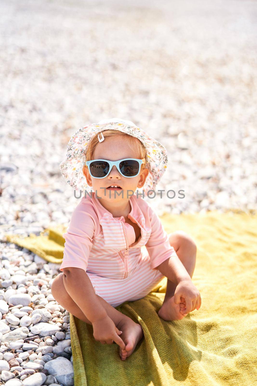 Little girl in sunglasses and panama sits on the beach by Nadtochiy