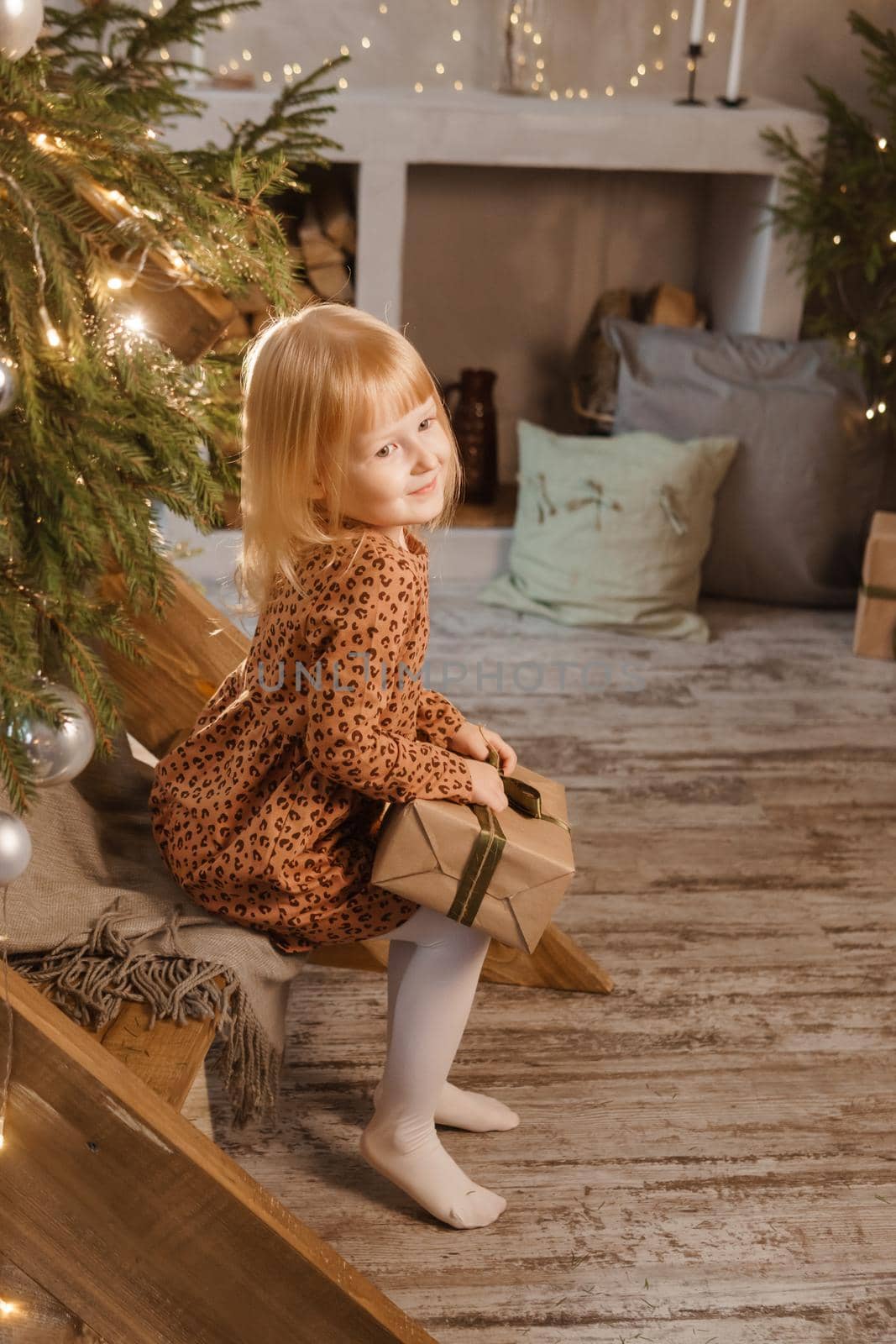 A little blonde girl is sitting on a wooden staircase in a Scandinavian interior decorated in a New Year's style. A child holds a Christmas gift in a craft package.