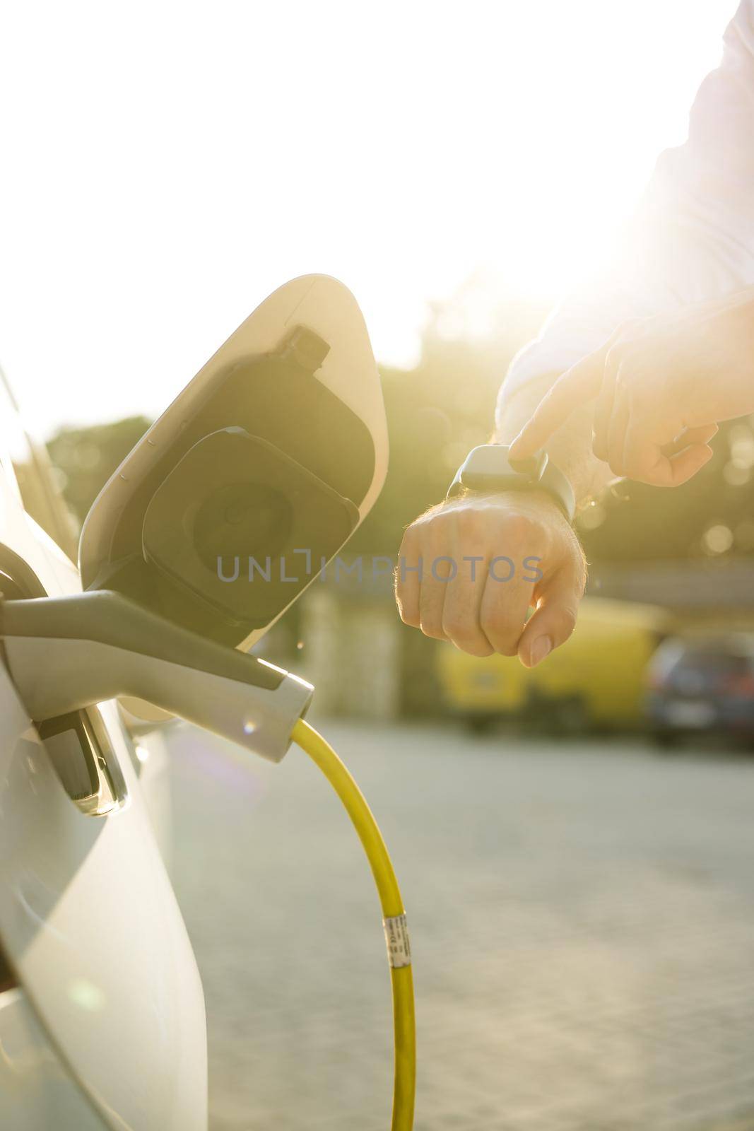 Unrecognizable man plugging in charging cable to to electric vehicle. Male hand inserts power connector into EV car and charges batteries uses smartwatch for activates start charging by uflypro