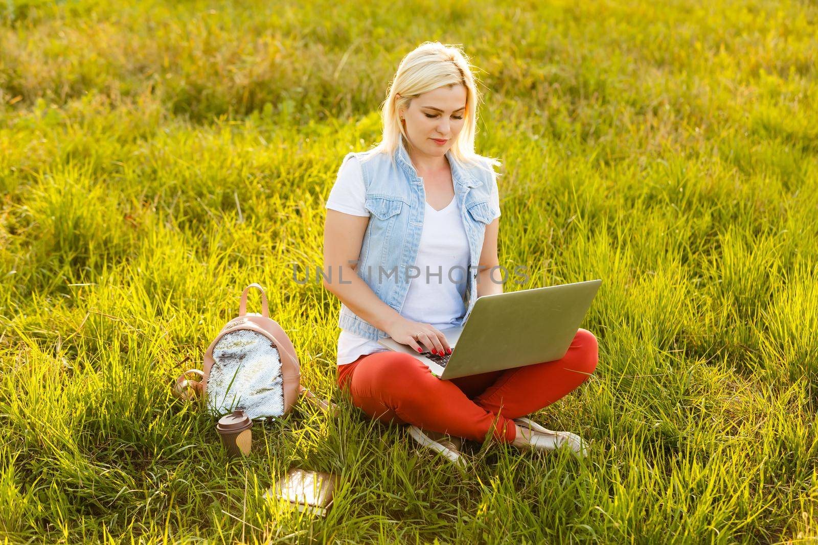 Portrait of pretty young woman sitting on green grass in park with legs crossed during summer day while using laptop for video call.
