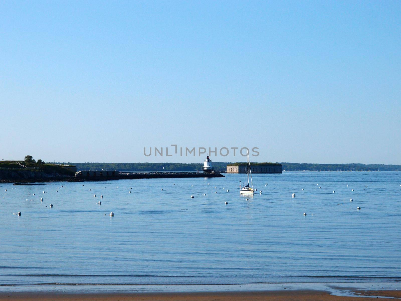 Beach with Portland Breakwater Lighthouse "Bug Light" at end of jetty rocky cove with boat and historic Fort Gorges visible on a beautiful day.