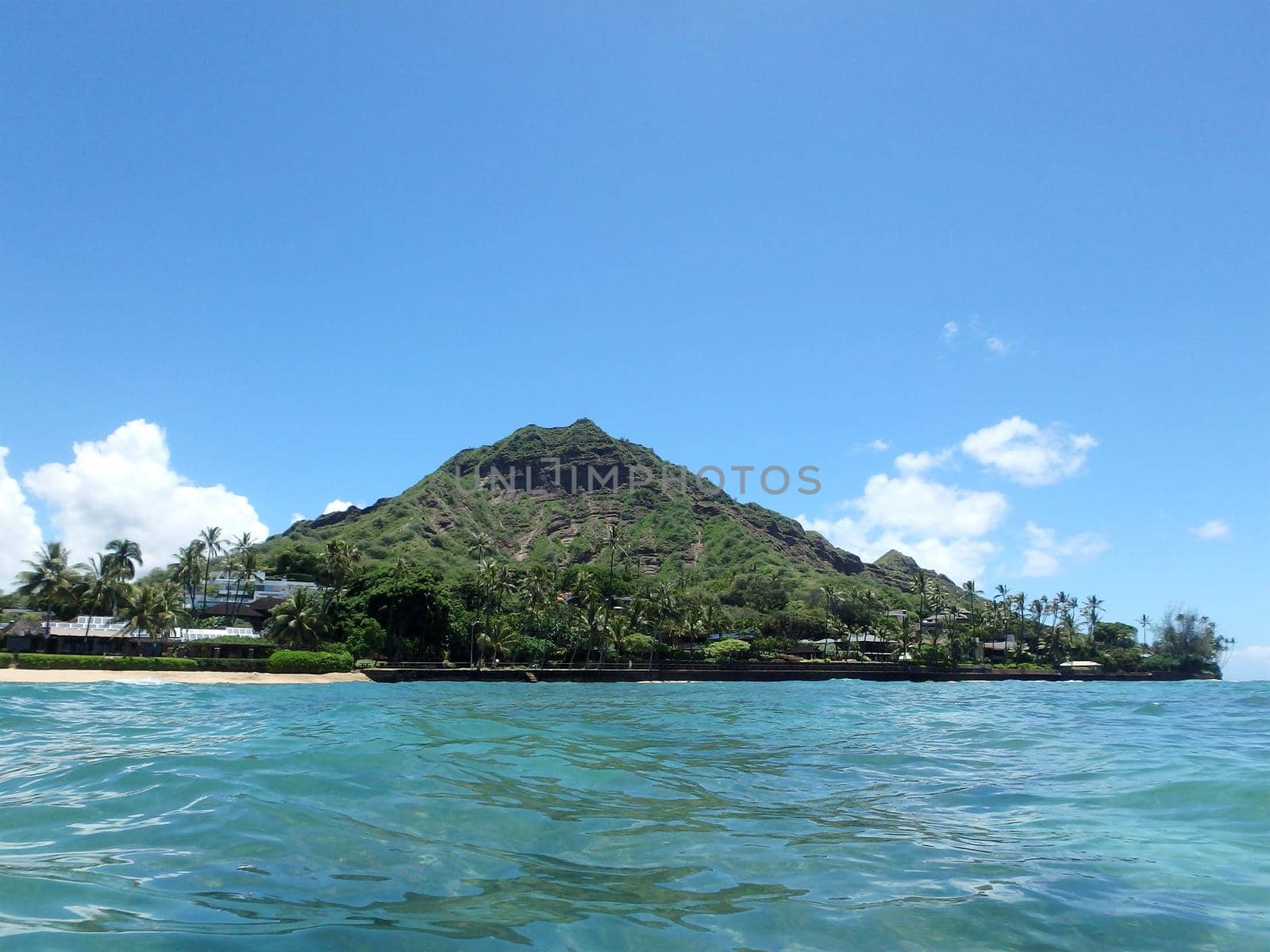 Beach and Makalei Beach Park with seawall, coconut trees, homes, and Diamond Head Crater in the distance on Oahu, Hawaii viewed from the water.