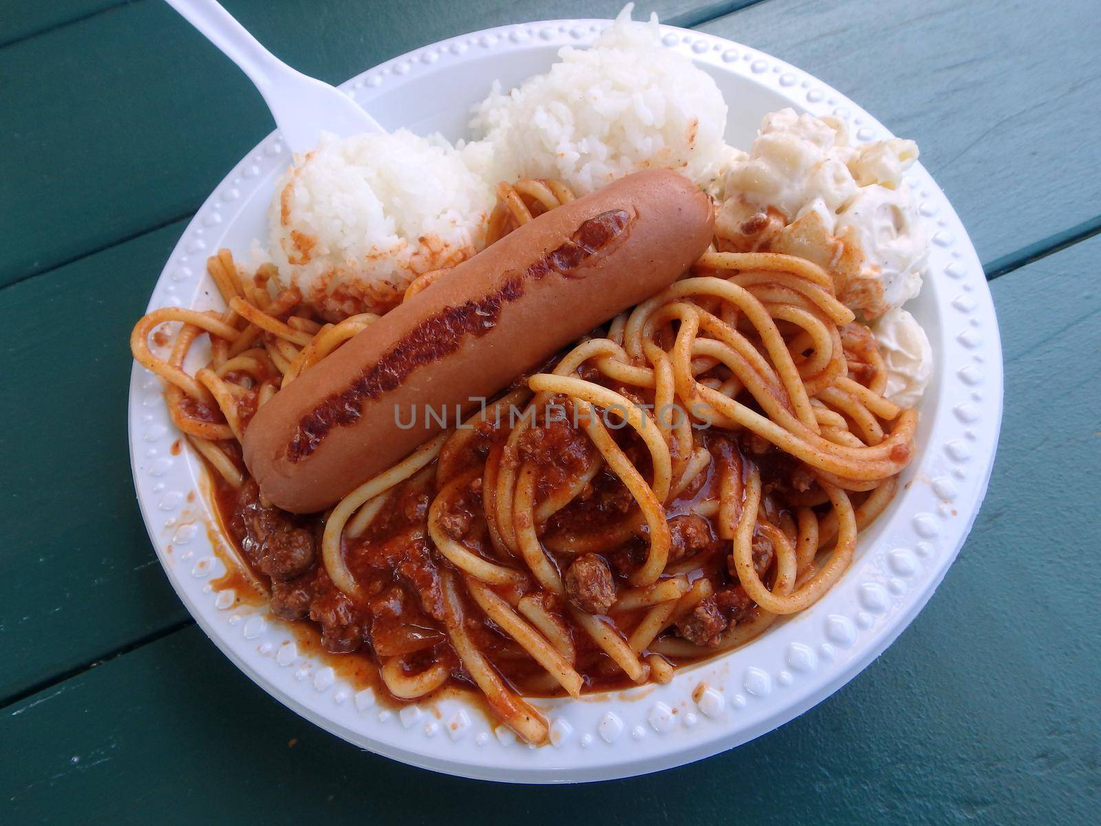 Spaghetti with meat sauce, Wiener hot dog, two scoops of rice, mac salad on a plastic plate with plastic fork on a green wood table.