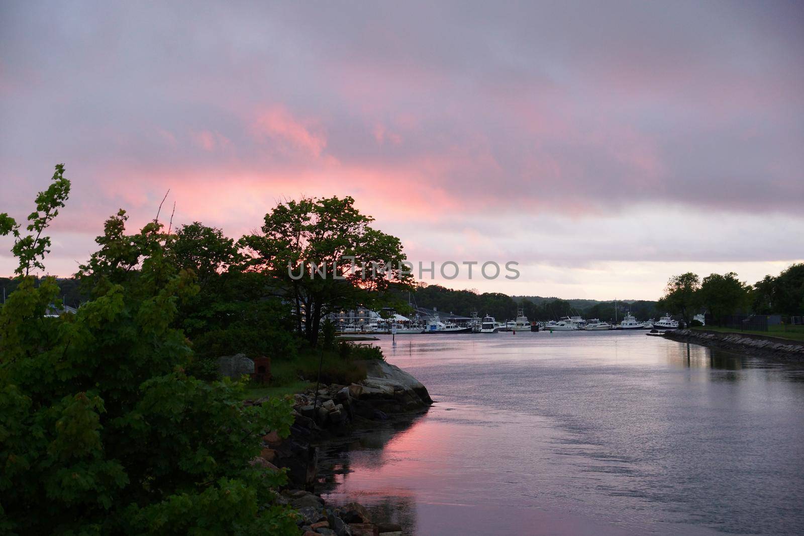 Sunset light shines over Blynman Canal reflects on the water with boats and buildings with trees behind them in in the distance Gloucester, Massachusetts.