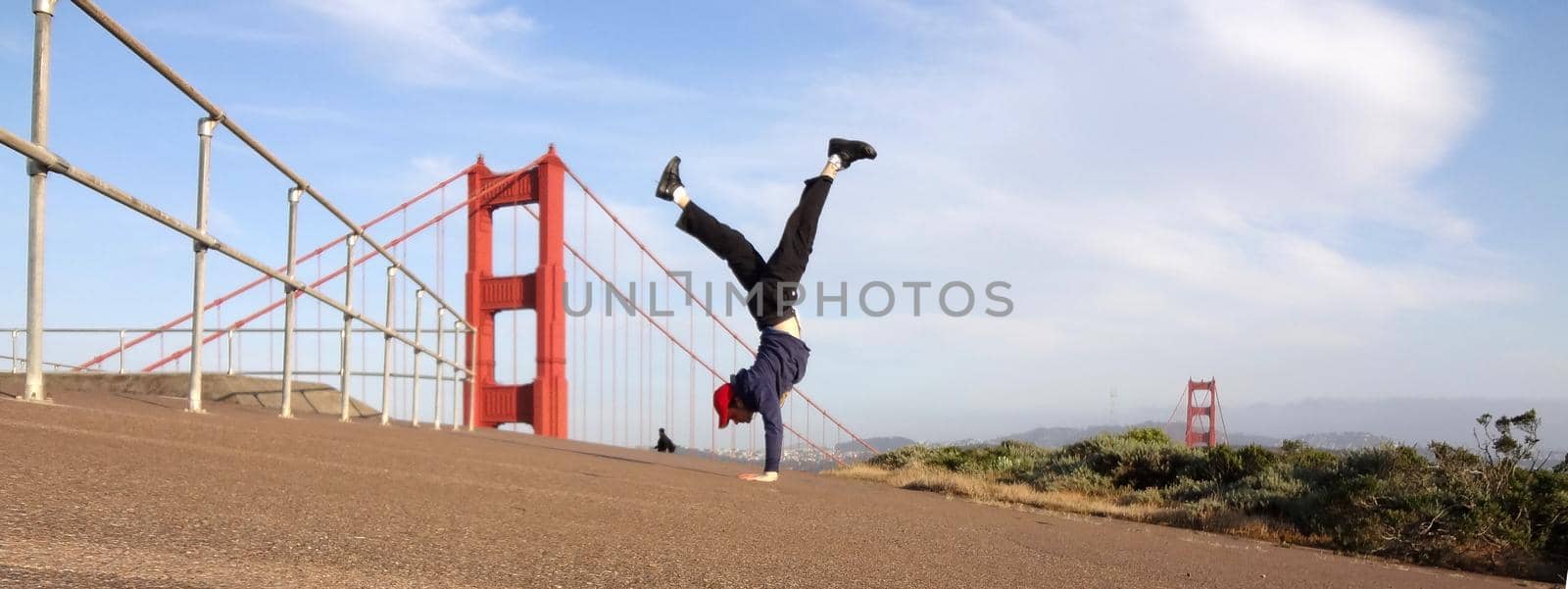 Man wearing hat, hoodie, long pants and shoes Handstands in front of the Golden Gate Bridge on the Marin side with San Francisco, California in the distance.