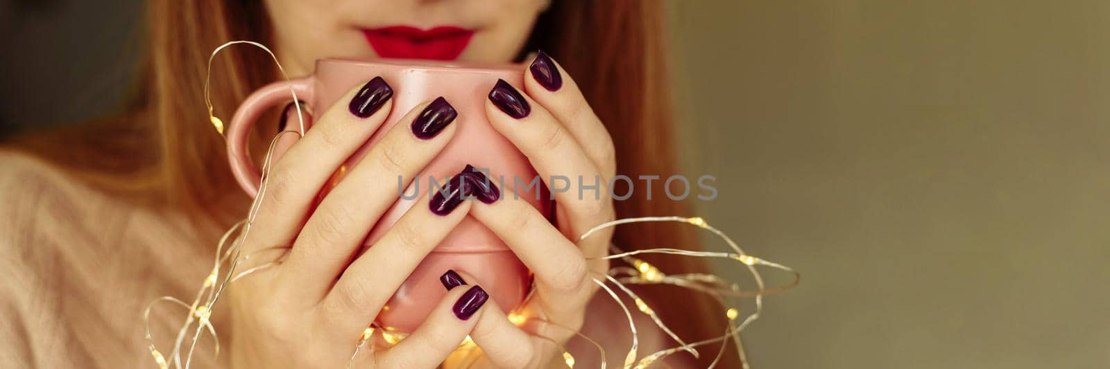 A young woman holds coffee near her mouth, drinks a warm drink in autumn or winter. Garland lights. Web banner, copy space.