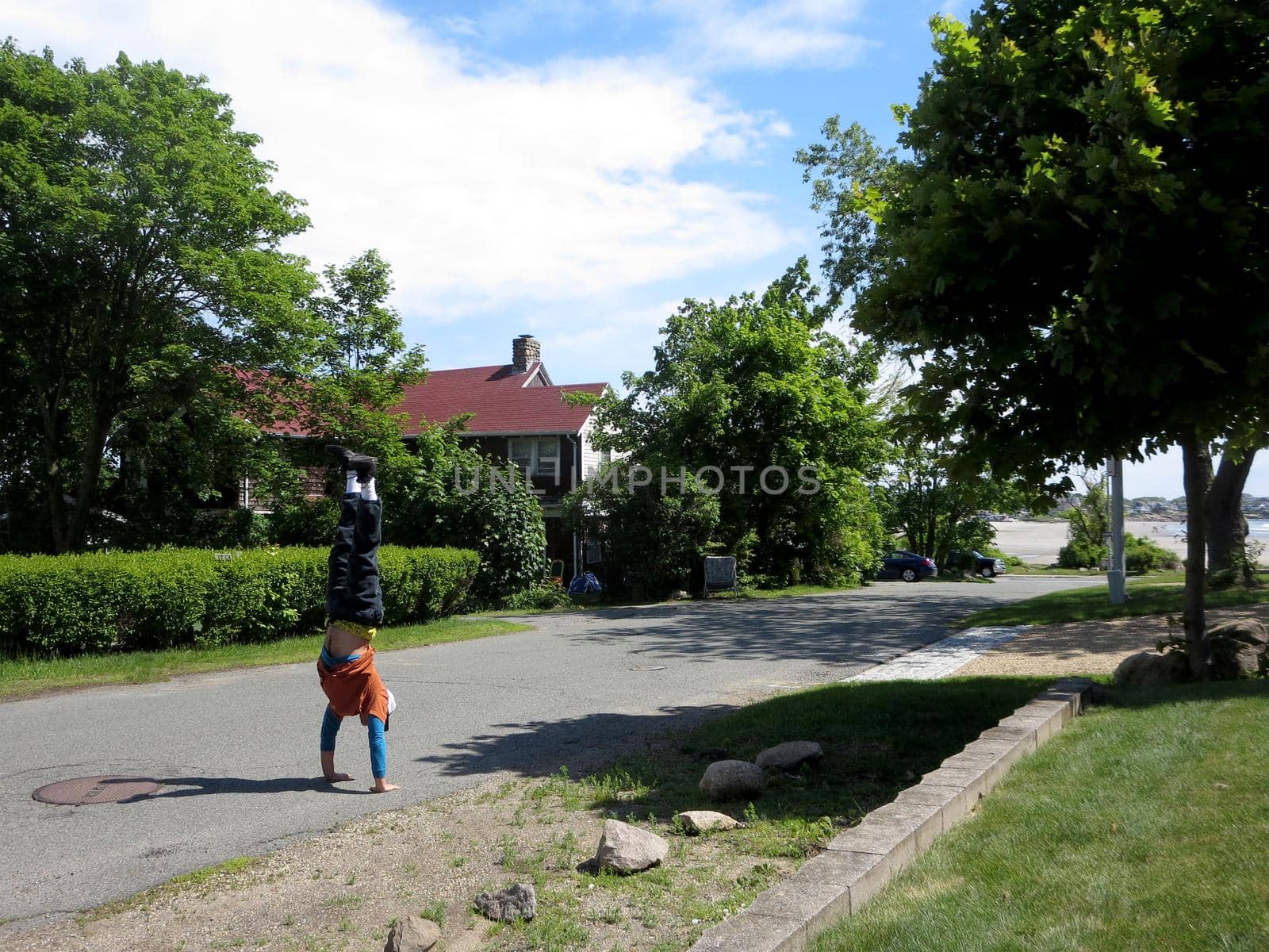 Man wearing a hat, t-shirt, long sleeve shirt, pants, and shoes Handstands on road of nice neighborhood near the beach which can be seen in the distance in Gloucester, Massachusetts