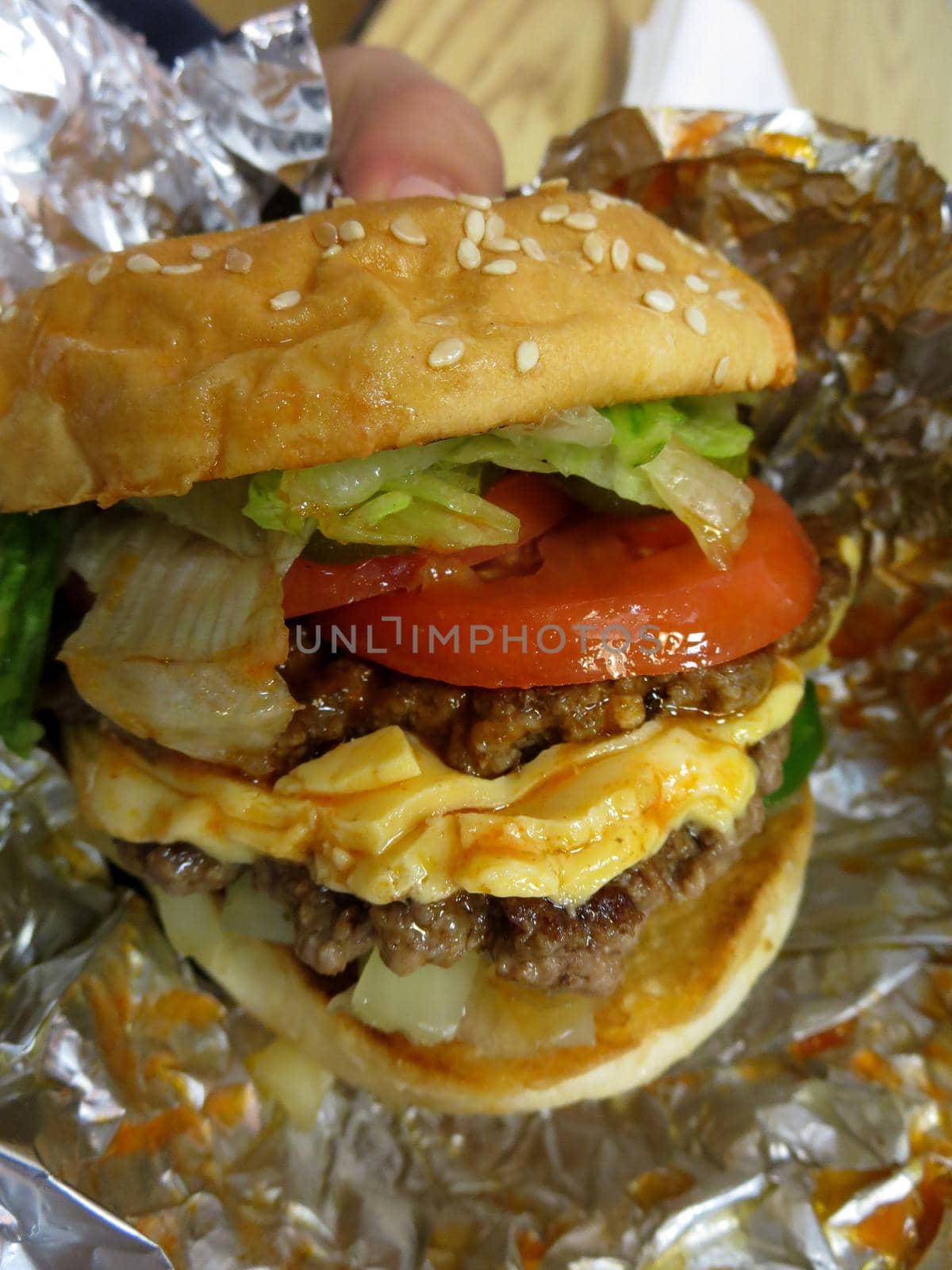 Close up of hands holding double cheeseburger with lettuce, tomatoes, and sauce wrapped in tinfoil.