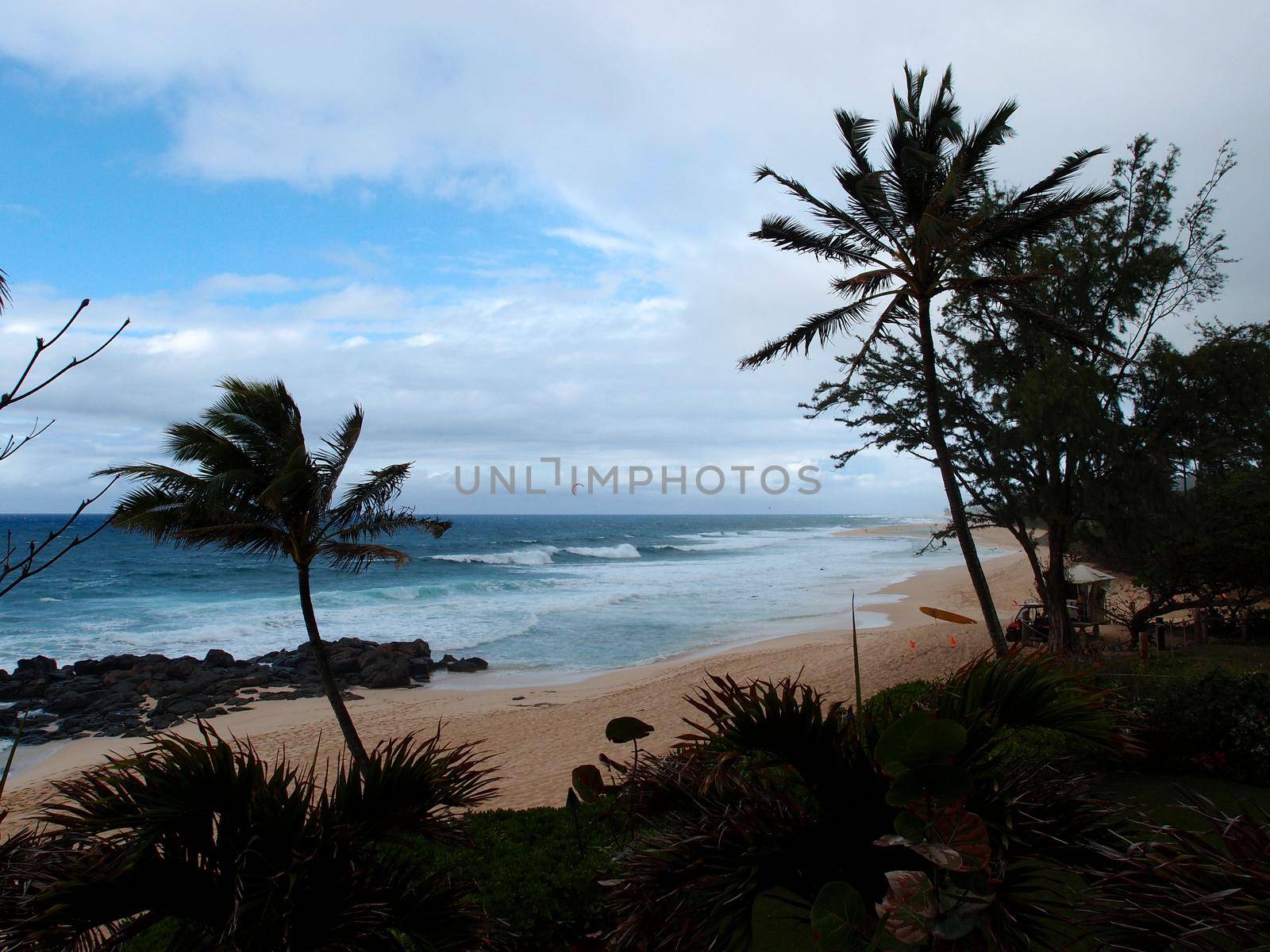 Waves lap on Beach at the world famous Banzai Pipeline with a lifeguard tower and  a kite surfer in the water on a cloudy day on Oahu, Hawaii.