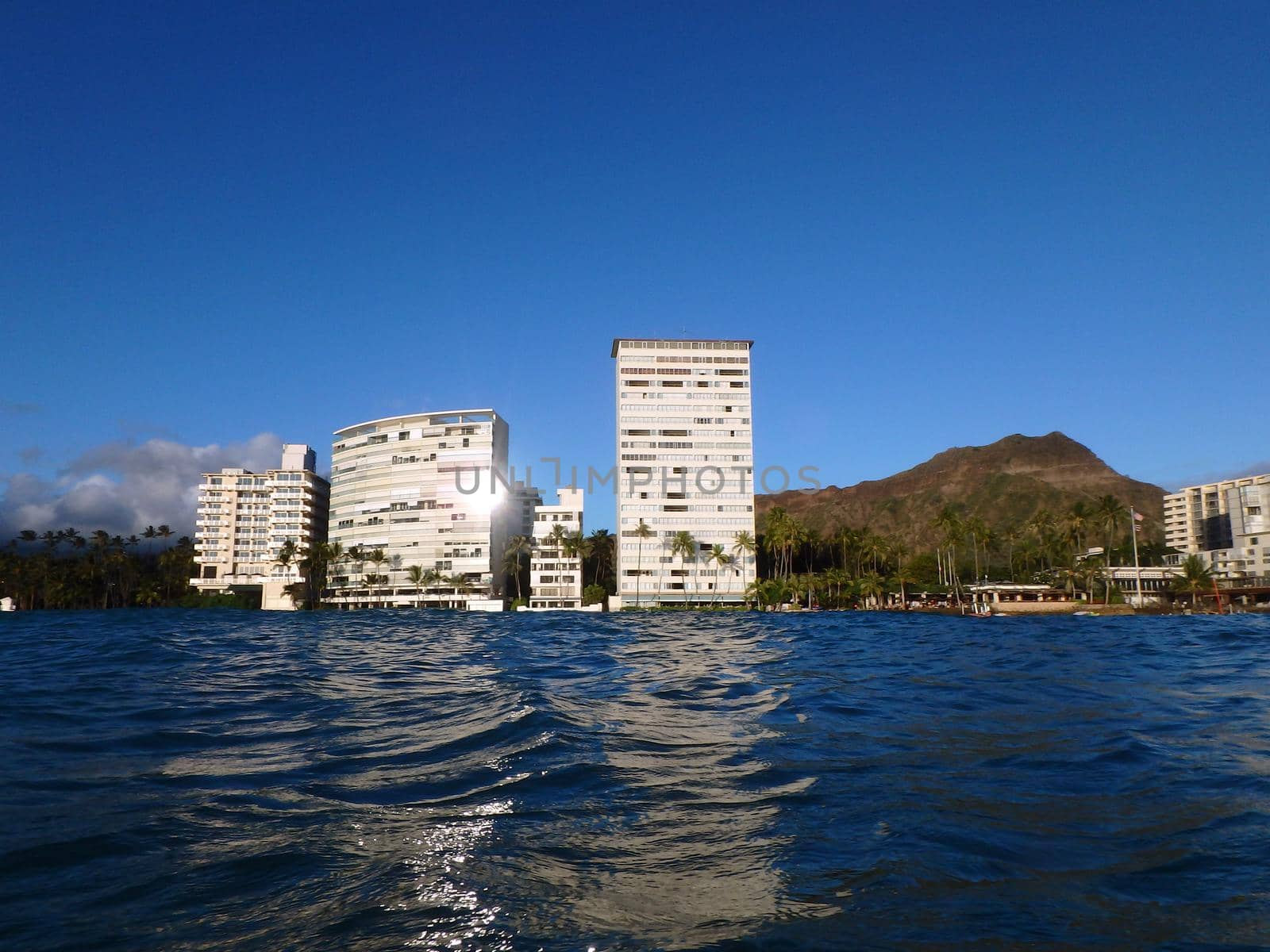 Sun reflects off of Hotel building as the hotel reflects off the water with coconut trees, Condo buildings, clouds and Diamond Head Crater in the distance by EricGBVD