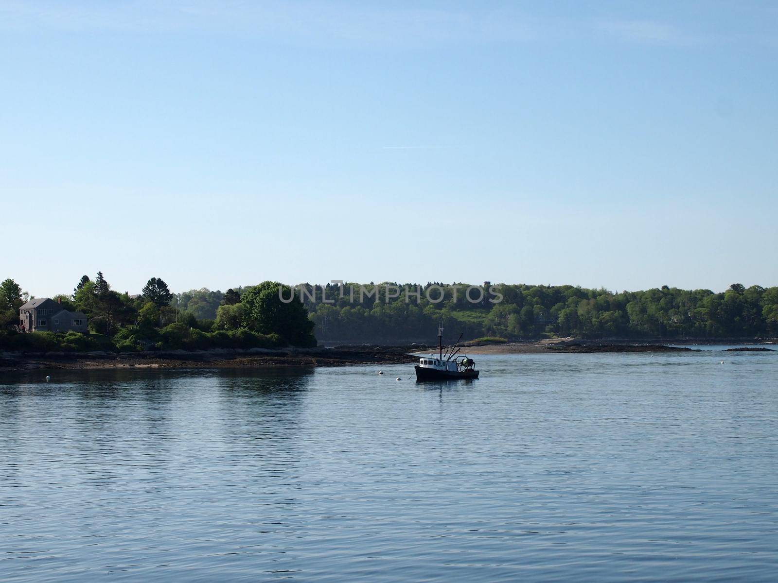 Fishing boat rests in water of Casco Bay off shore of island with house and trees in Maine.