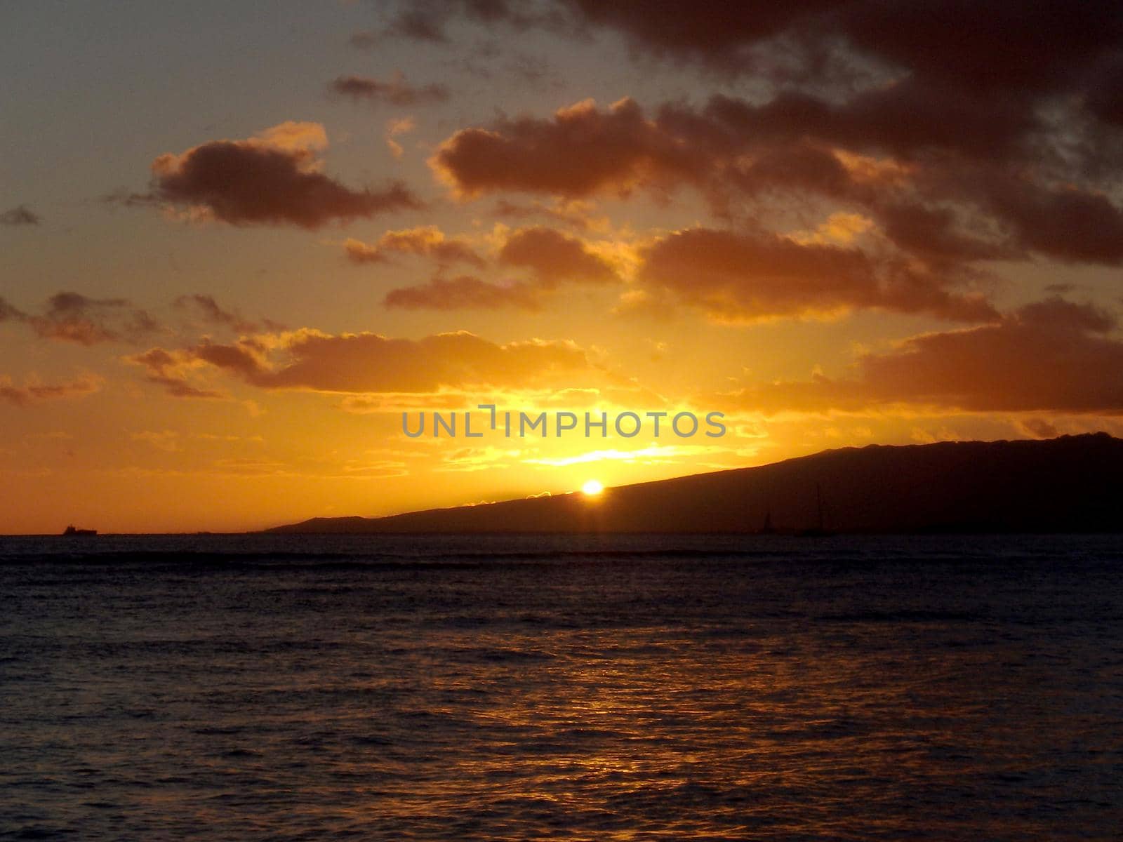 dramatic lighting as Sunsets behind Waianae mountains with light reflecting on ocean and illuminating the sky with boats sailing on the water off Waikiki on Oahu, Hawaii.