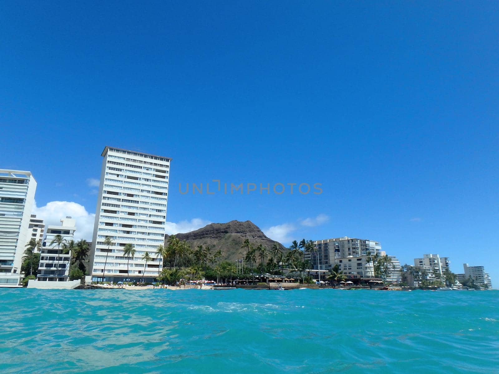 Hotel building, Outrigger Canoe Club, coconut trees, Condo buildings, clouds, and Diamond Head Crater in the distance on Oahu, Hawaii viewed from the water on a beautiful day. 