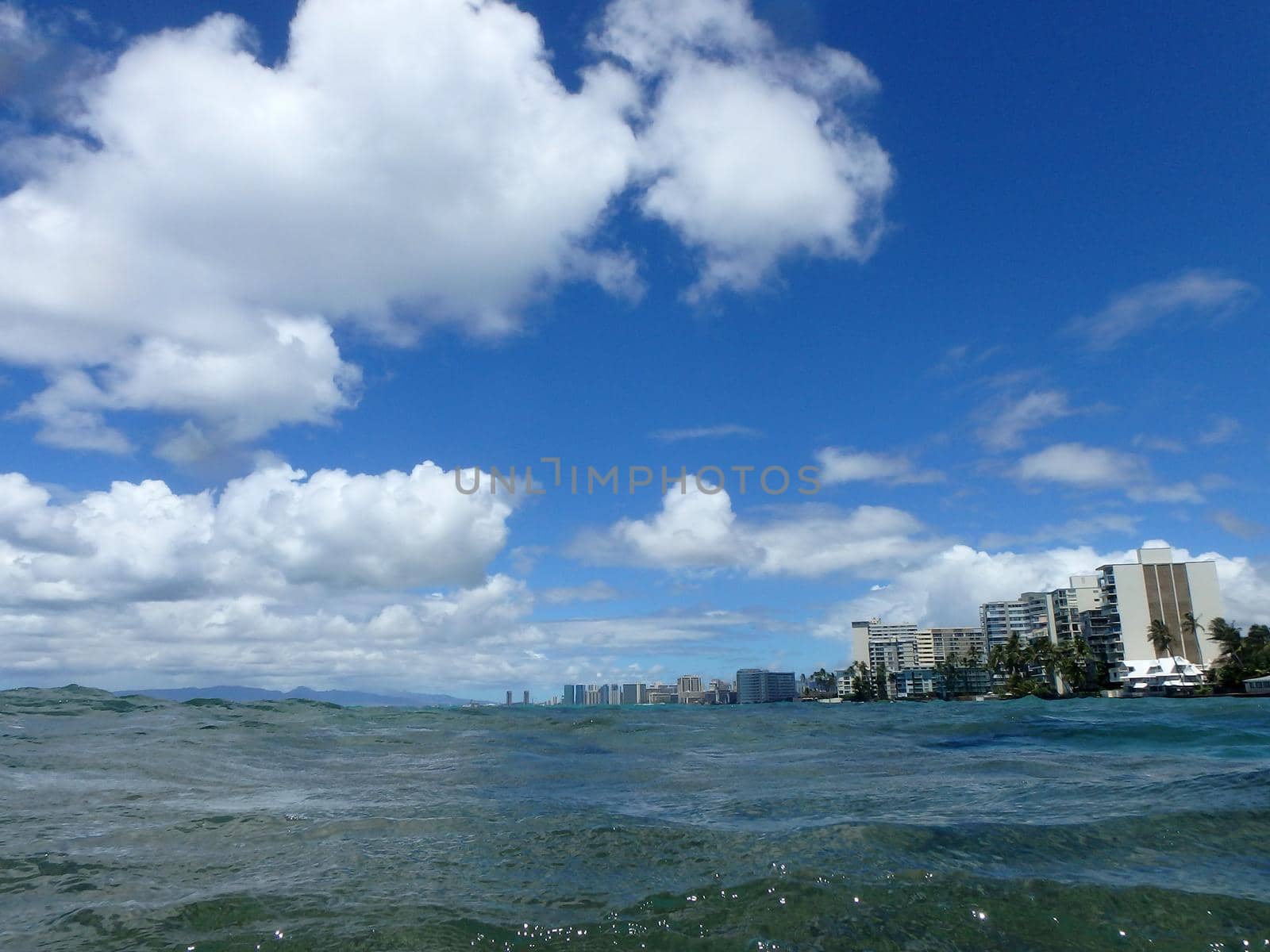 Ocean waters ripple off the coast of waikiki with hotels lining the water and clouds in the sky on Oahu.